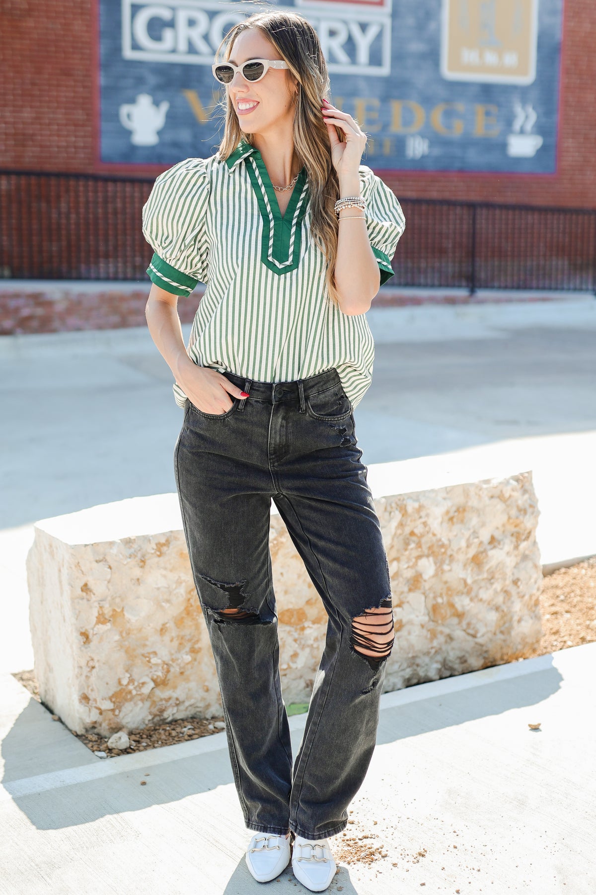 Woman wearing a green and white striped shirt with puffed sleeves and ripped black jeans, standing in front of a store sign.