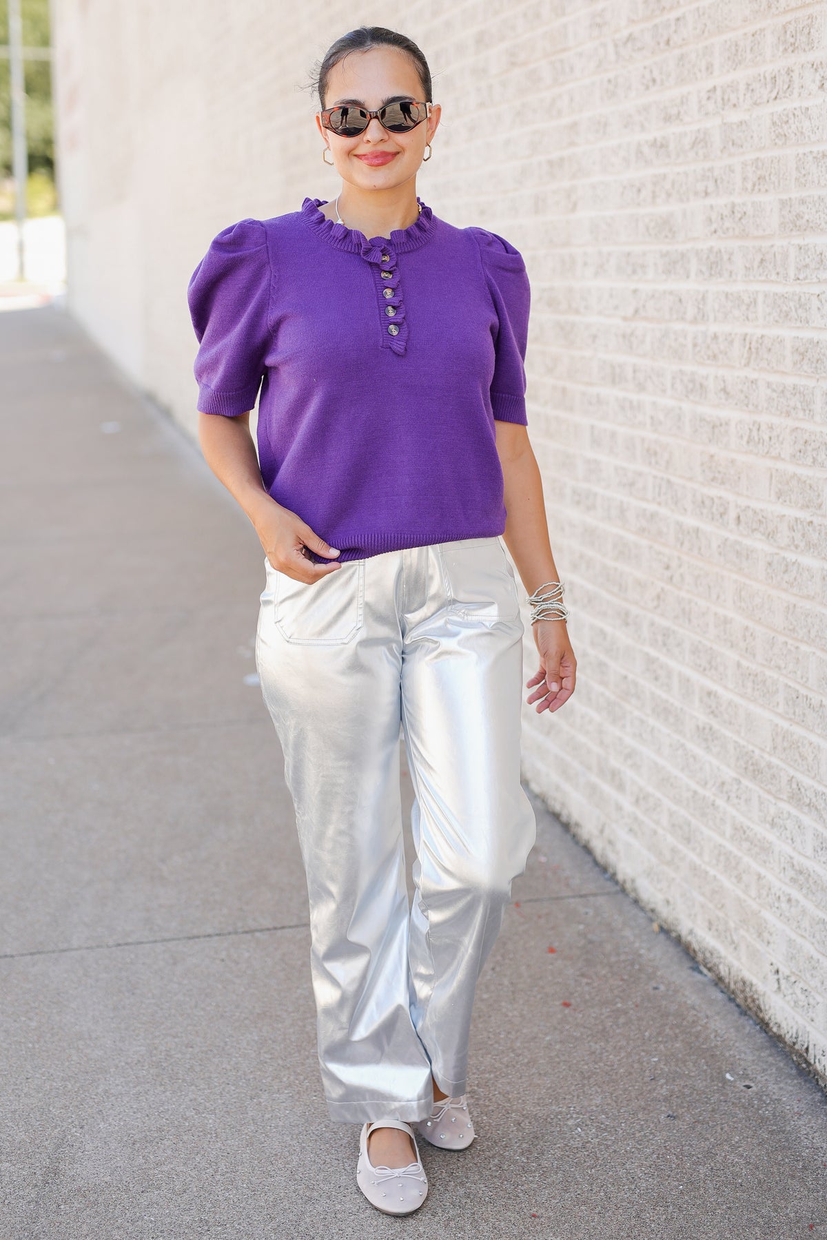A woman wearing a purple short-sleeve knit top with puff sleeves and a buttoned ruffled neckline, paired with shiny metallic silver pants, walking outdoors next to a light brick wall.