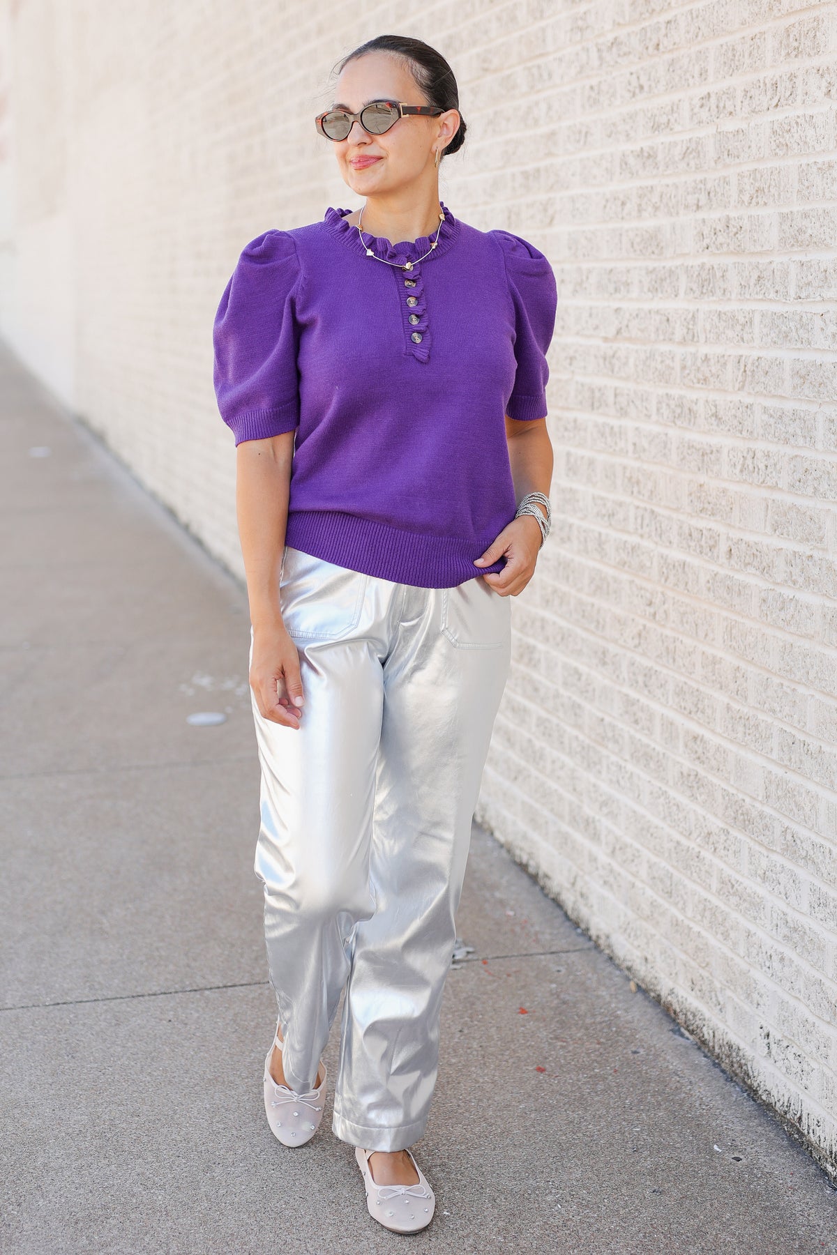 A woman wearing a purple puff-sleeve top with a buttoned neckline, shiny silver pants, and white flats, standing on a sidewalk next to a white brick wall.