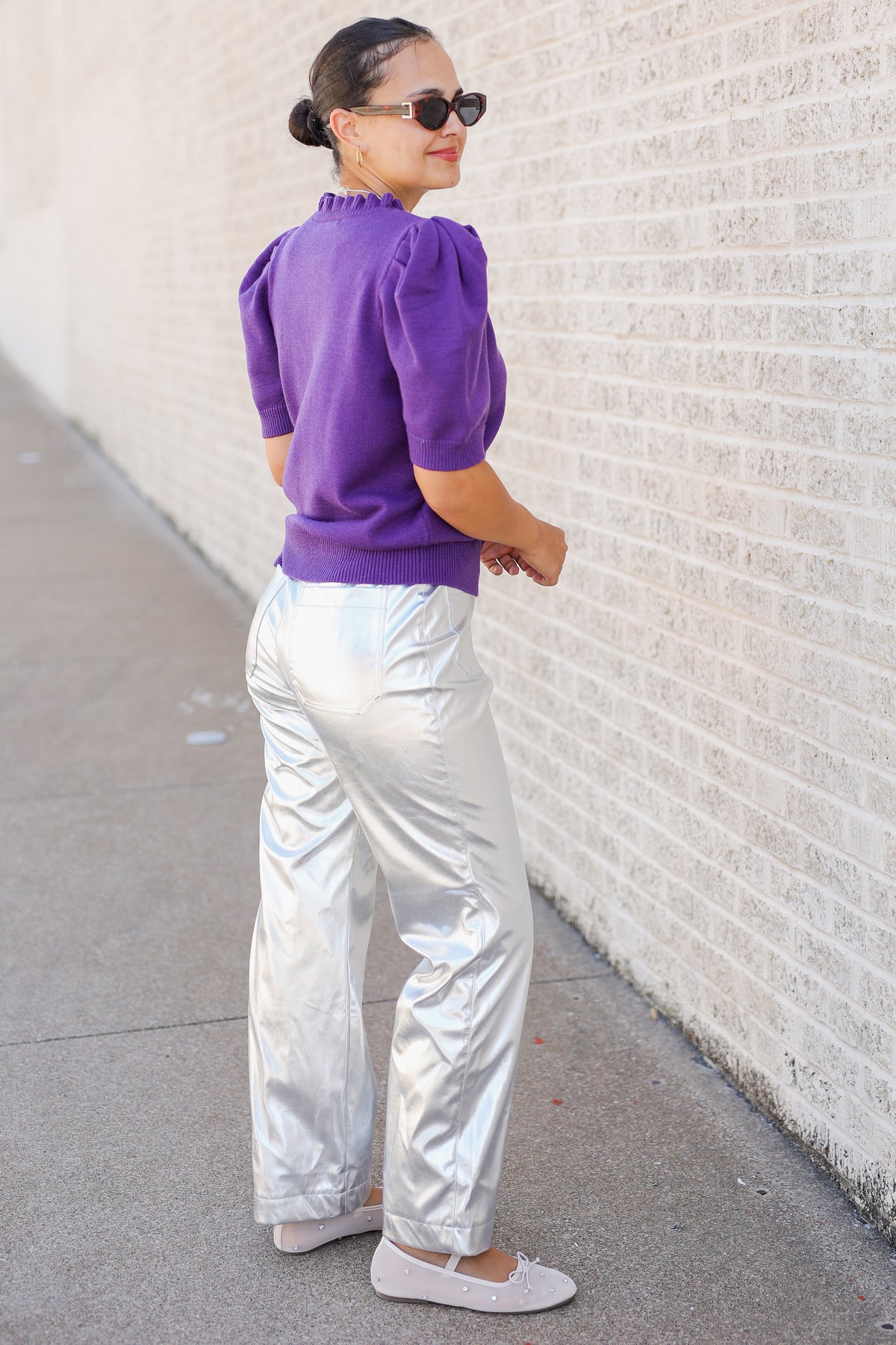 A woman wearing a purple puff-sleeve top and shiny silver pants stands on a sidewalk next to a white brick wall, looking over her shoulder.