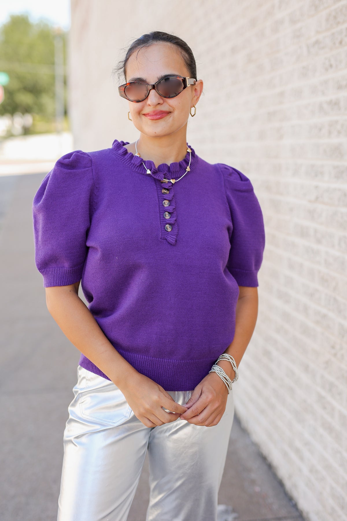 A woman wearing a vibrant purple short-sleeve knit top with puff sleeves, a ruffled collar, and decorative buttons, paired with shiny silver pants, standing outdoors against a light brick wall.