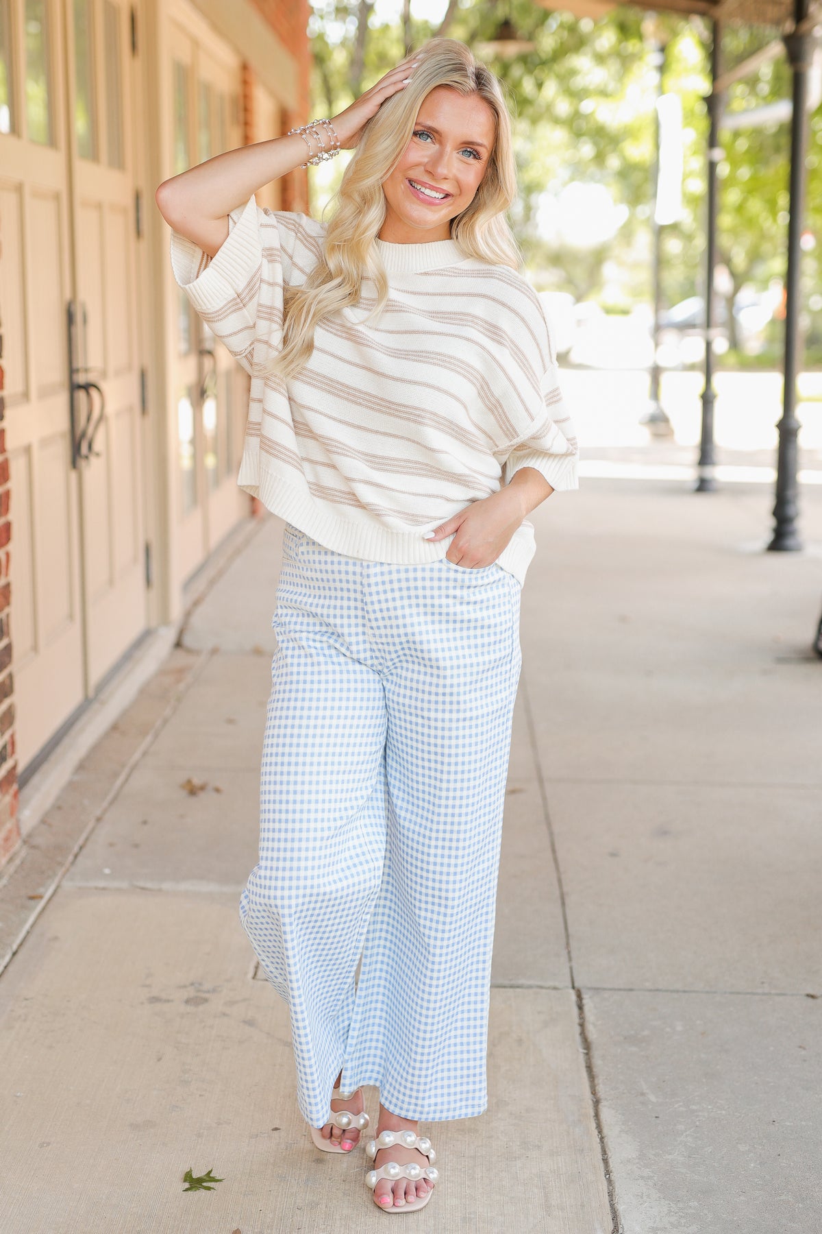Blonde model wearing a taupe knit top and blue gingham pants, standing outside.