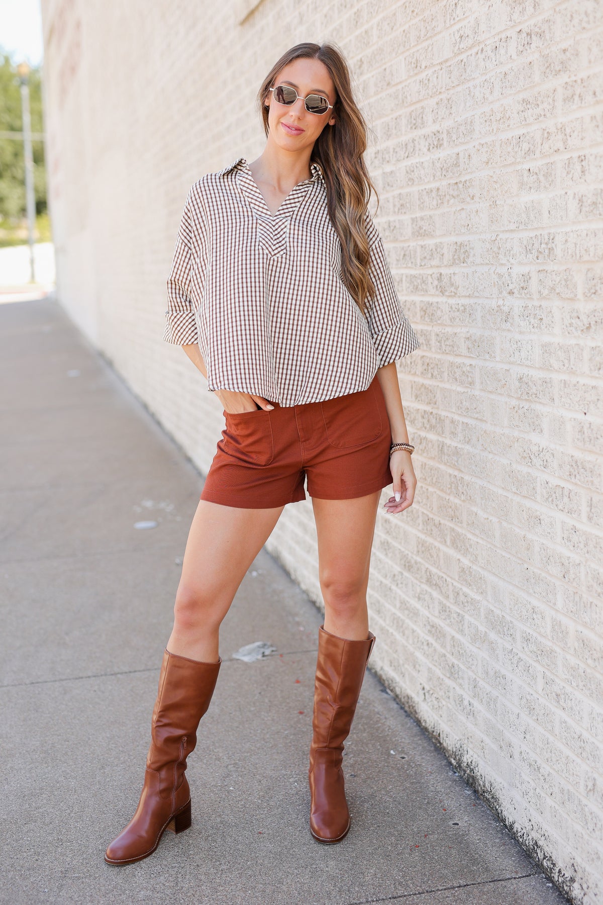 A woman stands outdoors against a light brick wall wearing a beige and white gingham button-up shirt, rust-brown shorts, and brown knee-high boots.