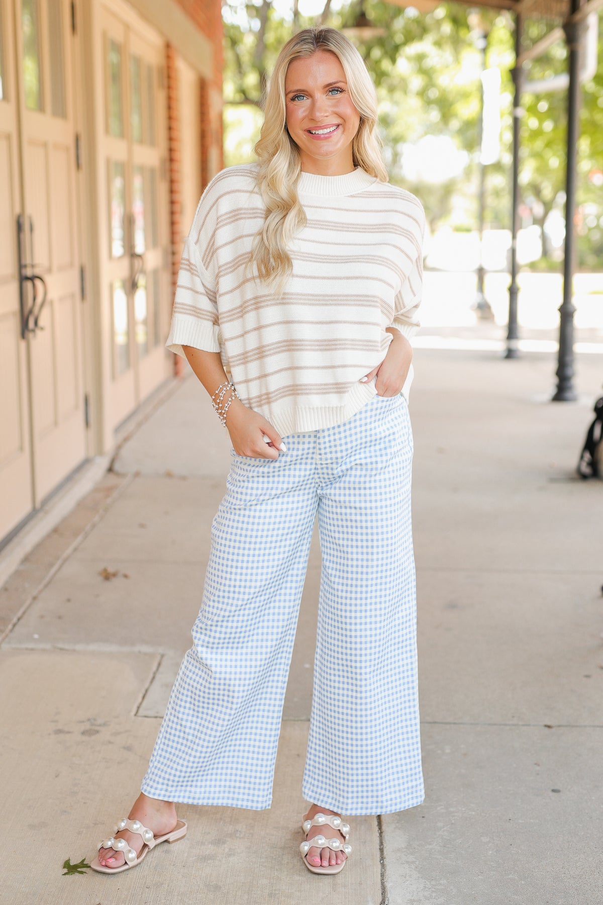 Blonde model wearing a taupe knit top and blue gingham pants, standing outside.