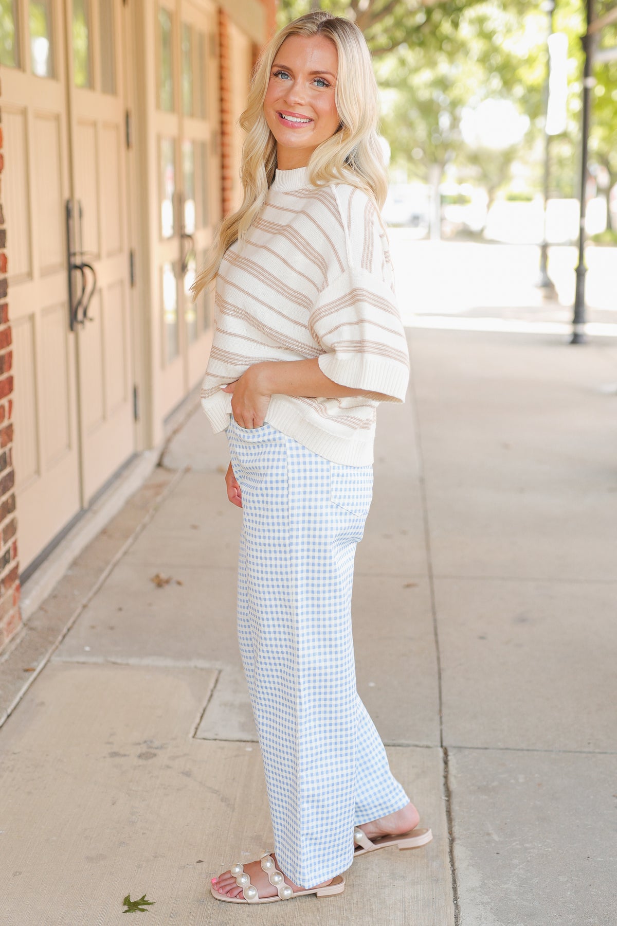 Blonde model wearing a taupe knit top and blue gingham pants, standing outside.