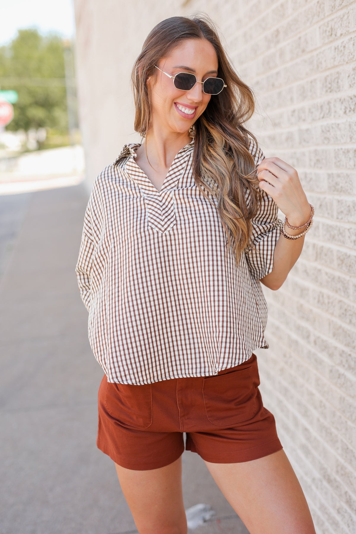 A woman wearing a brown and white gingham button-down shirt and rust-brown shorts, posing outdoors against a light brick wall.