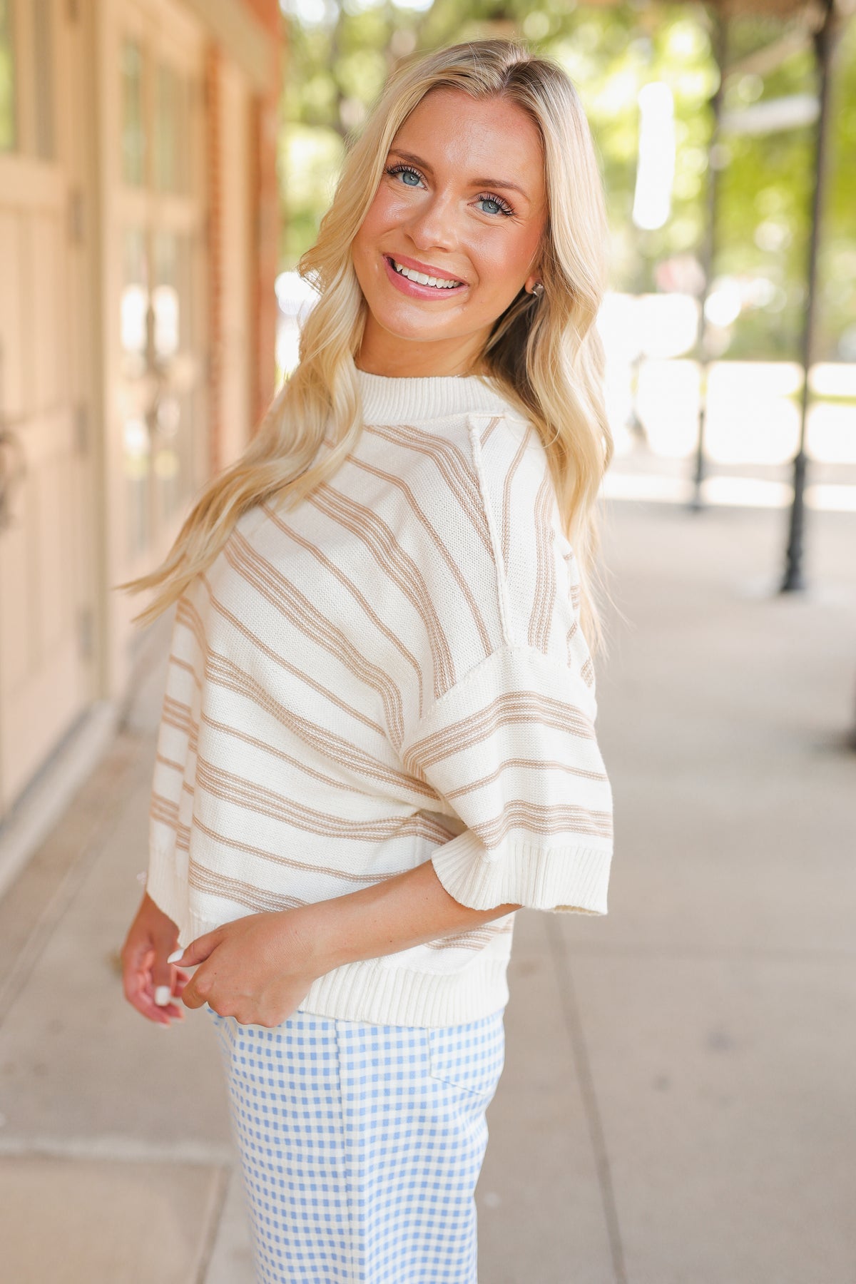 Blonde model wearing a taupe knit top and blue gingham pants, standing outside.
