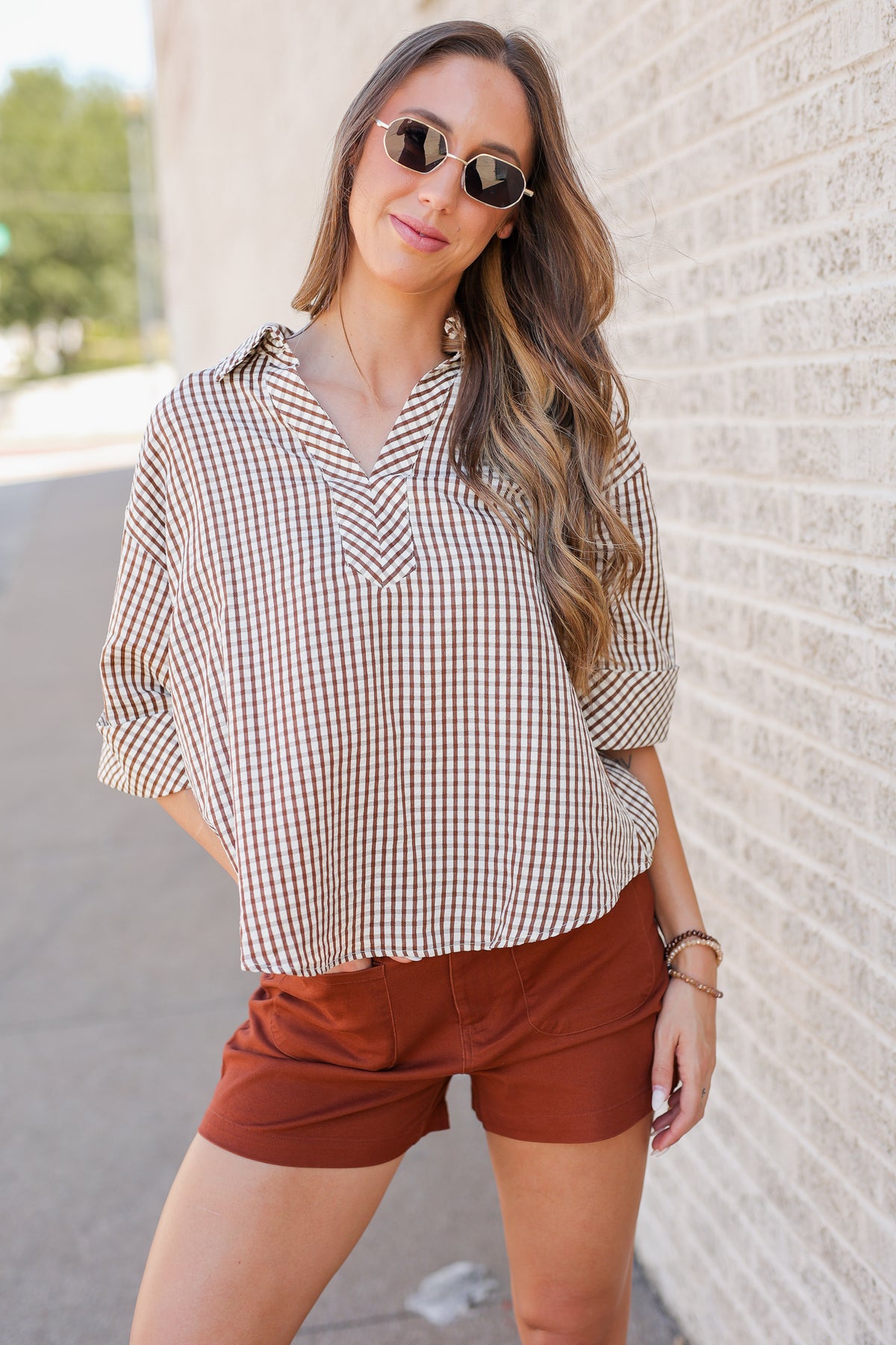 A woman wearing a brown and white gingham button-down shirt with rolled sleeves and rust-brown shorts, standing outdoors against a light brick wall.