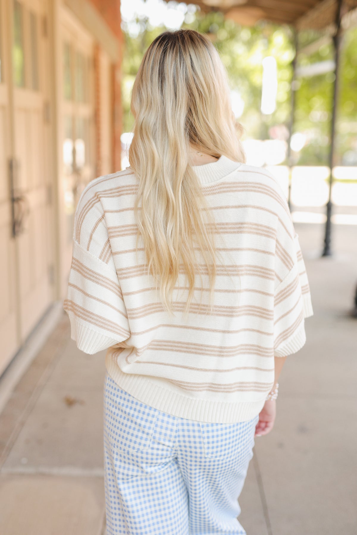 Blonde model wearing a taupe knit top and blue gingham pants, standing outside.