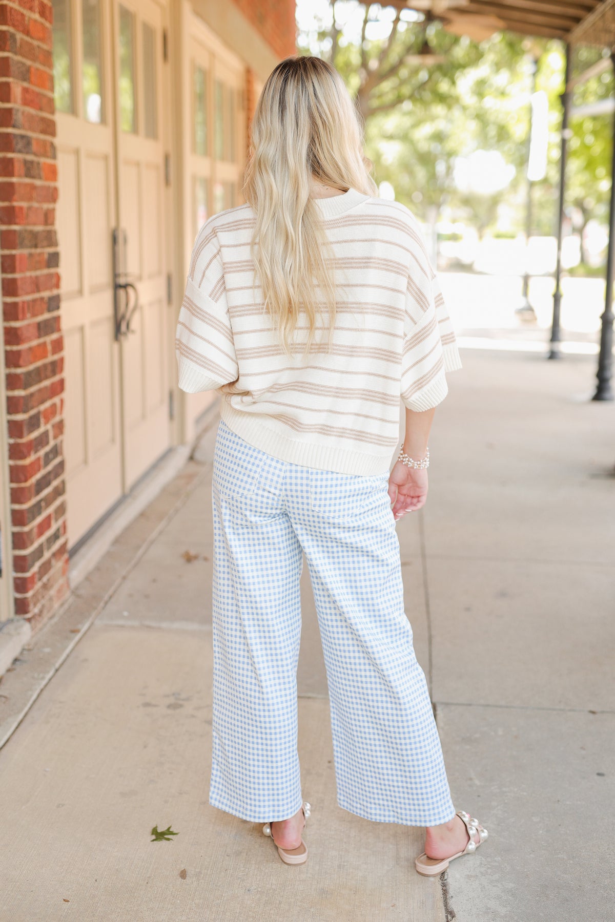 Blonde model wearing a taupe knit top and blue gingham pants, standing outside.