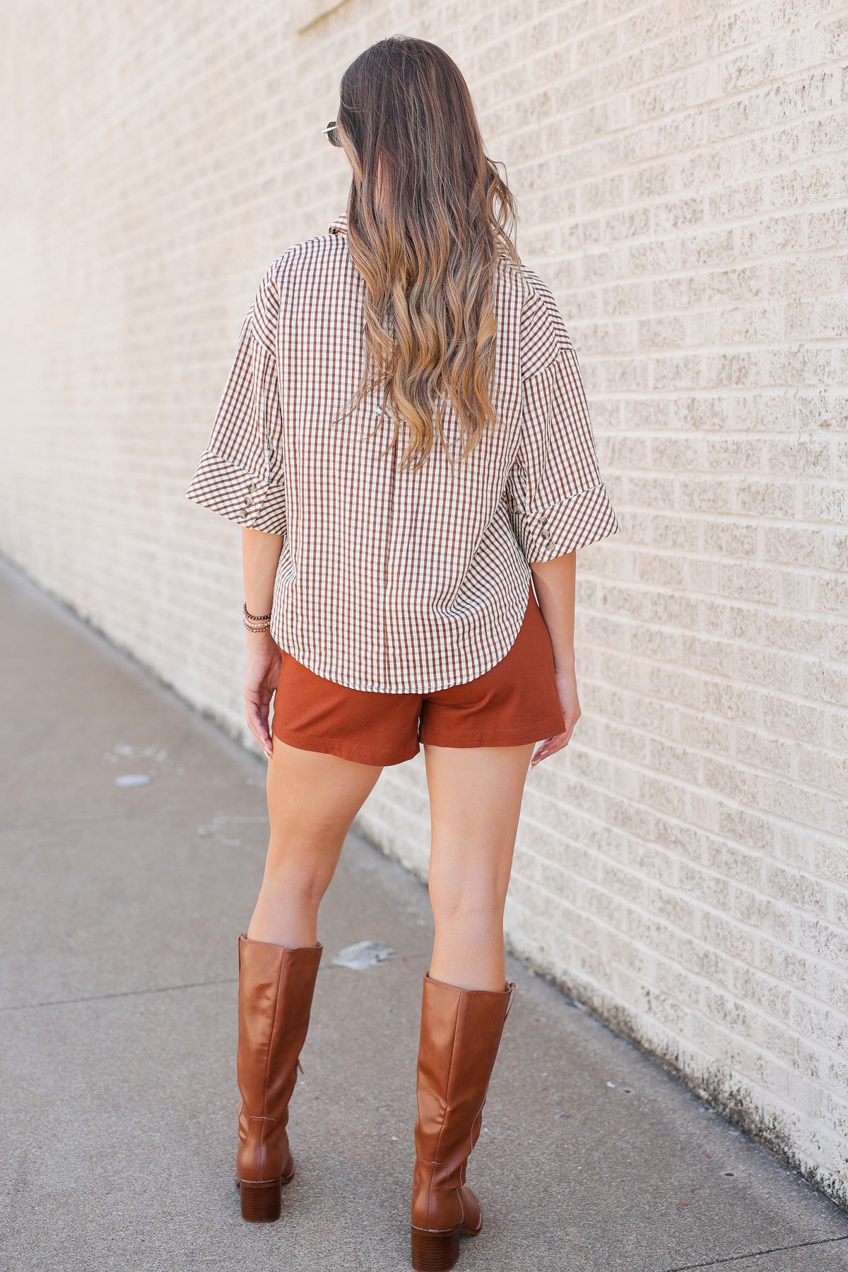 A woman photographed from behind wearing a brown and white gingham check shirt, rust-colored shorts, and brown knee-high boots, standing against a light-colored brick wall.