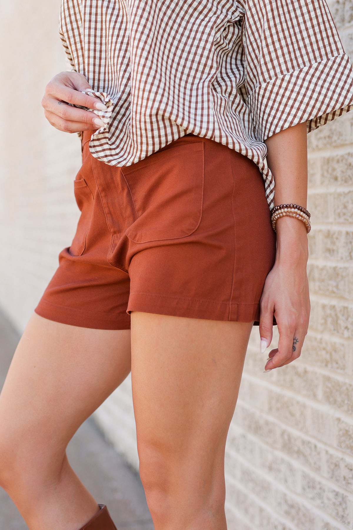 A person wearing rust-orange shorts and a brown-and-white gingham blouse, standing against a light stone wall.
