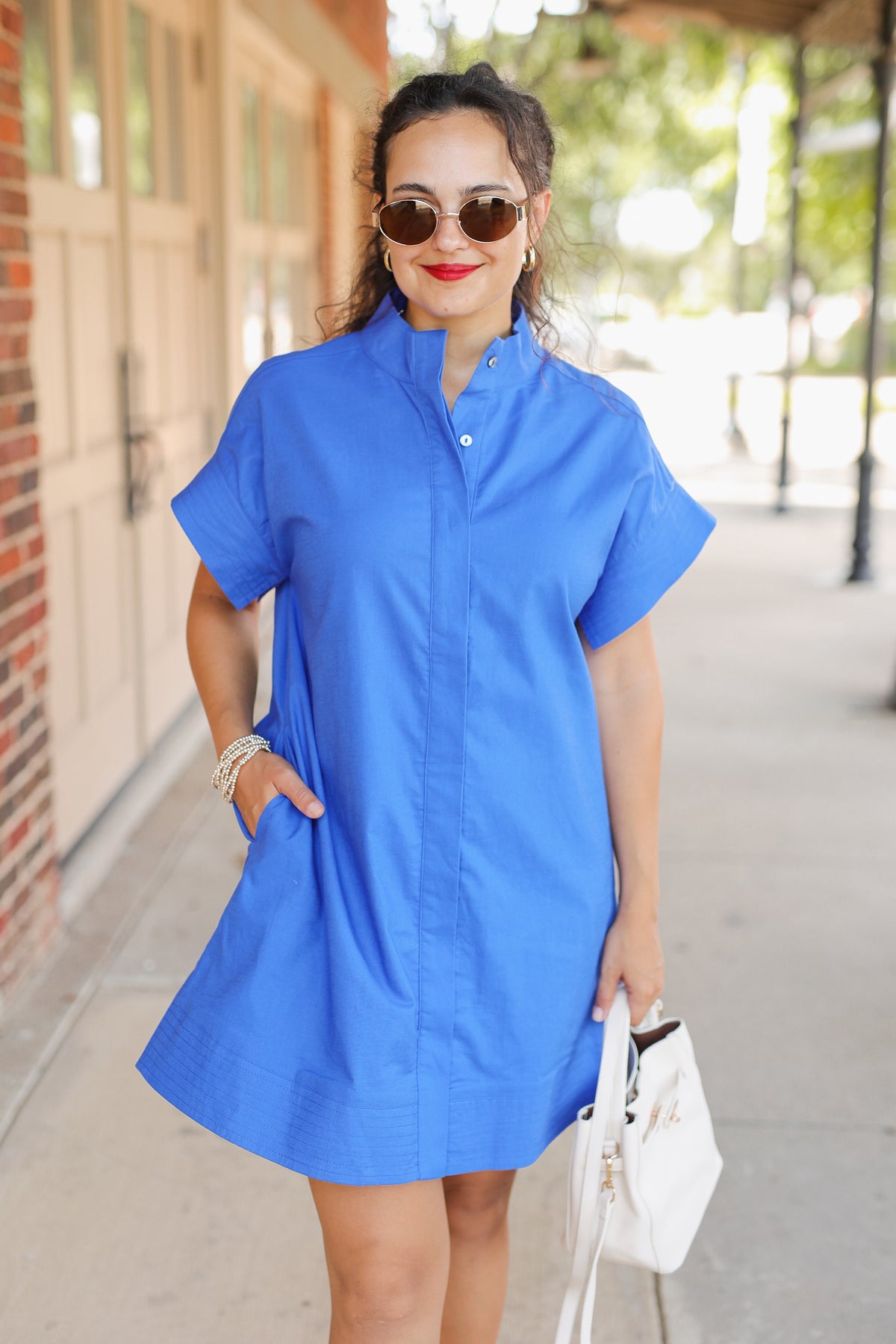 Woman wearing a blue dress with sunglasses and a white handbag on a sidewalk.