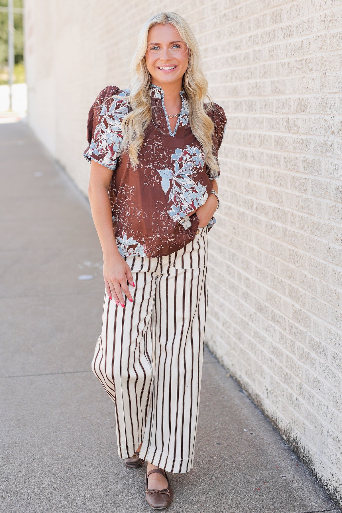 A woman stands on a sidewalk next to a light brick wall, wearing a brown short-sleeve blouse with a white floral print and cream-and-brown vertically striped wide-leg pants.