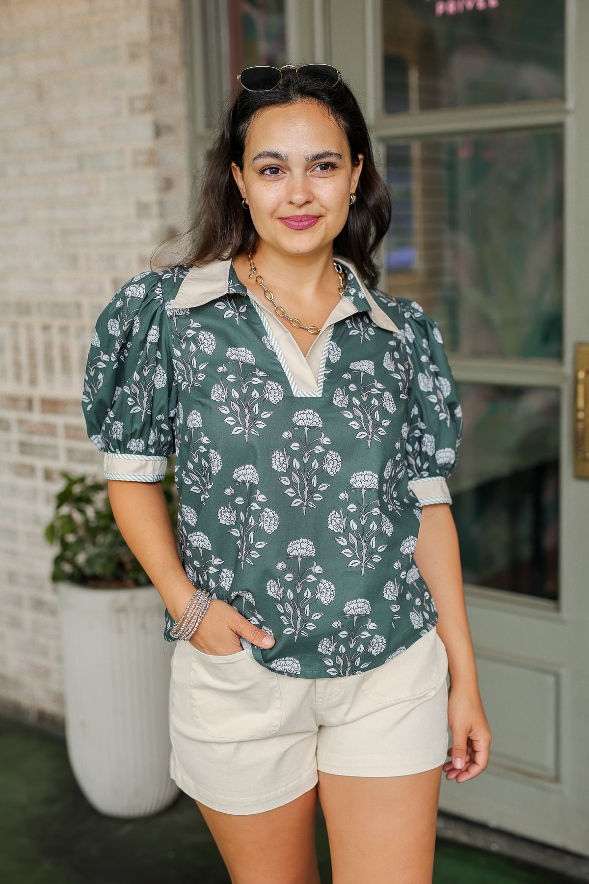 Woman wearing a green floral blouse and white shorts standing outdoors.