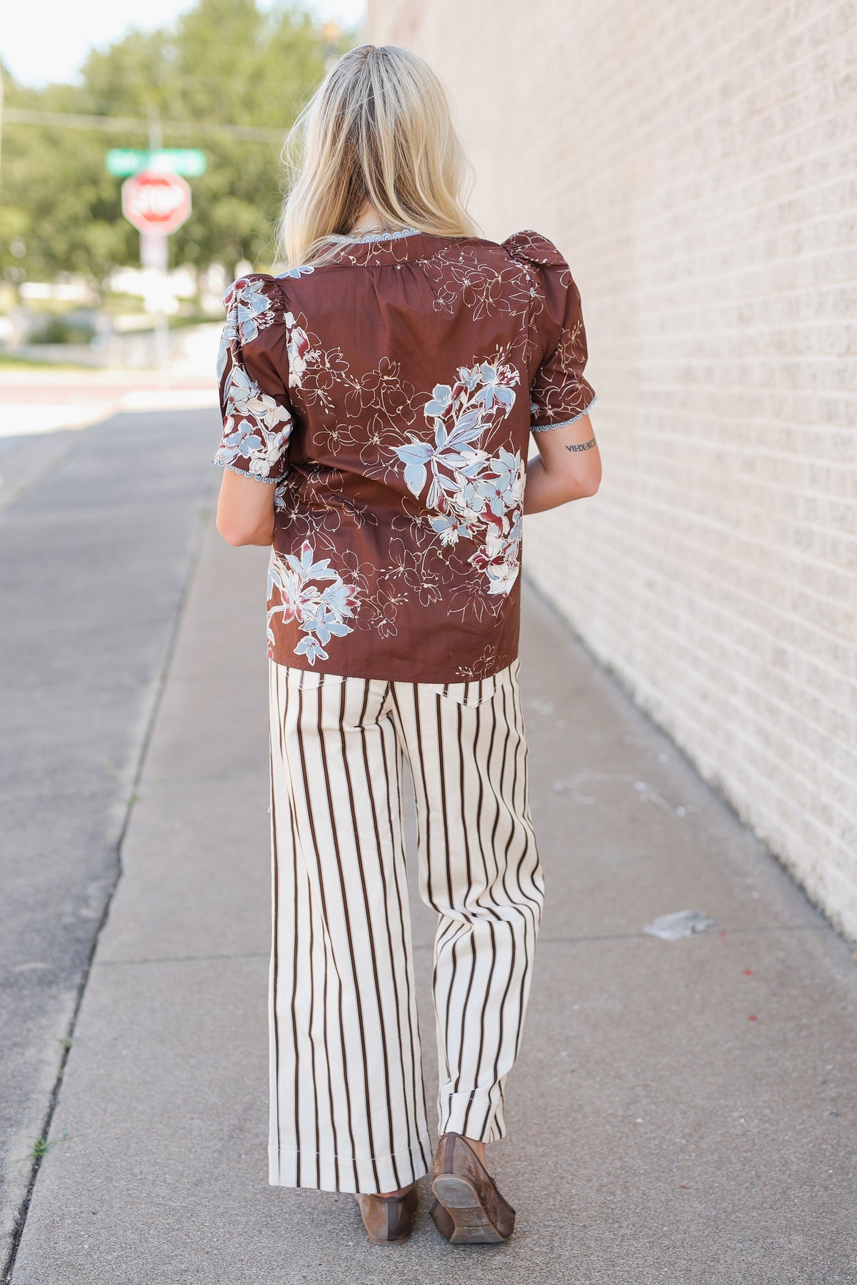 A woman viewed from behind wearing a brown short-sleeve blouse with a white floral pattern and cream-and-brown vertically striped wide-leg pants, standing on a sidewalk next to a light brick wall.
