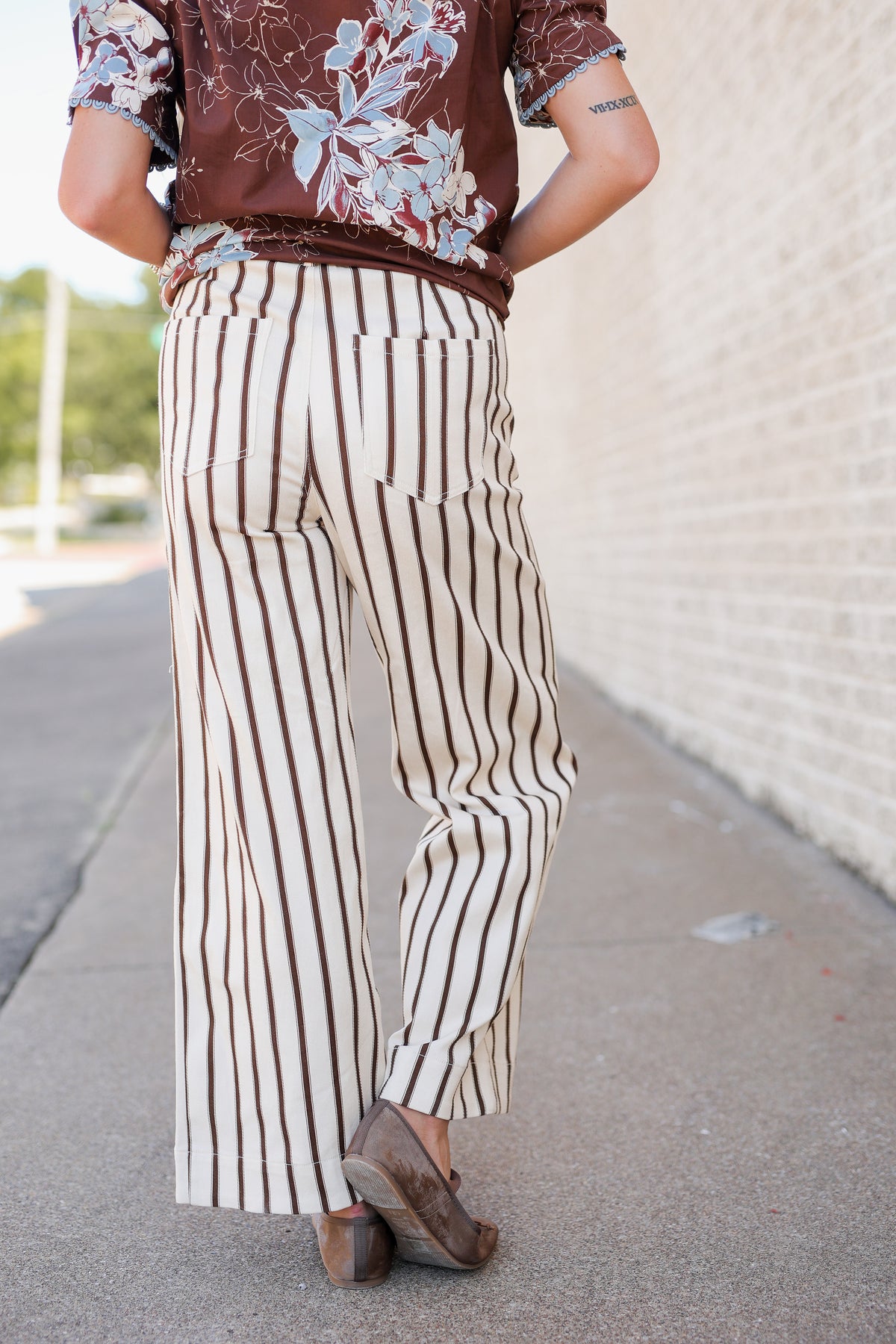A person shown from the back wearing cream wide-leg pants with vertical brown stripes, a brown floral short-sleeve top, and brown wedge shoes, standing outdoors on a sidewalk next to a light brick wall.