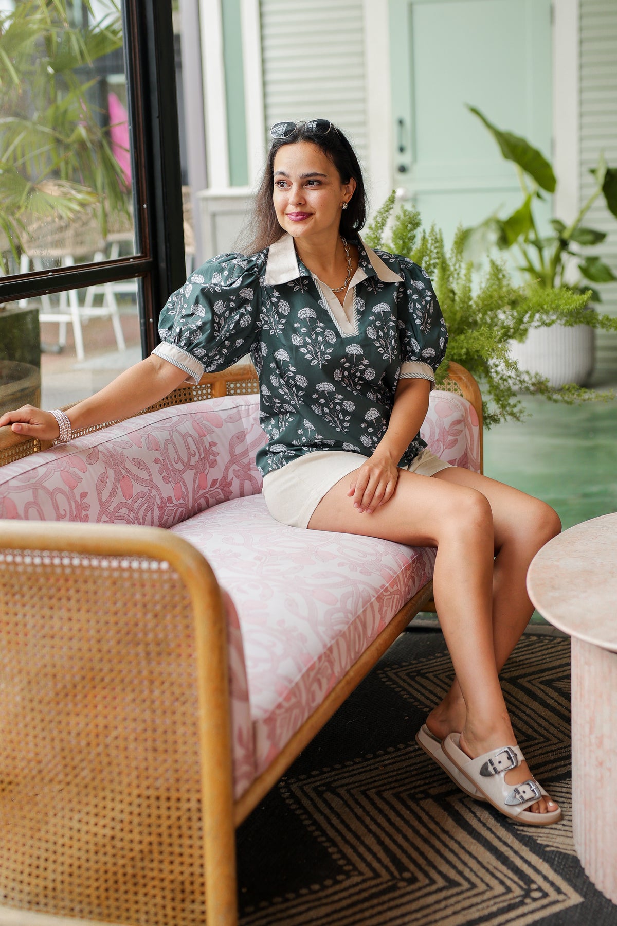 Woman sitting on a pink couch in a stylish outdoor setting with plants and a table.