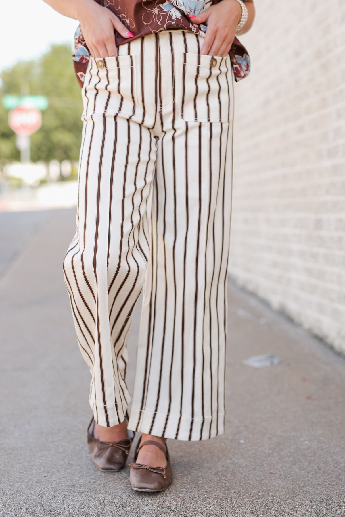 A person wearing cream wide-leg pants with vertical brown stripes, paired with a patterned top and brown flats, standing outdoors on a sidewalk next to a white brick wall.