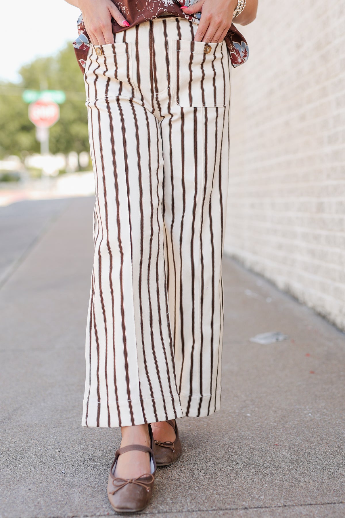 A person outdoors wearing wide-leg cropped pants with vertical brown and cream stripes, paired with brown flats, standing on a sidewalk beside a light brick wall.