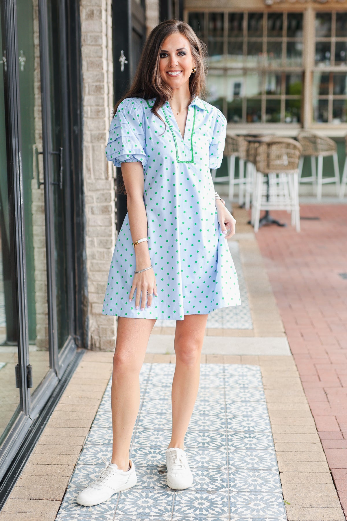 Woman wearing a light blue dress with white polka dots and white sneakers, standing on a sidewalk.