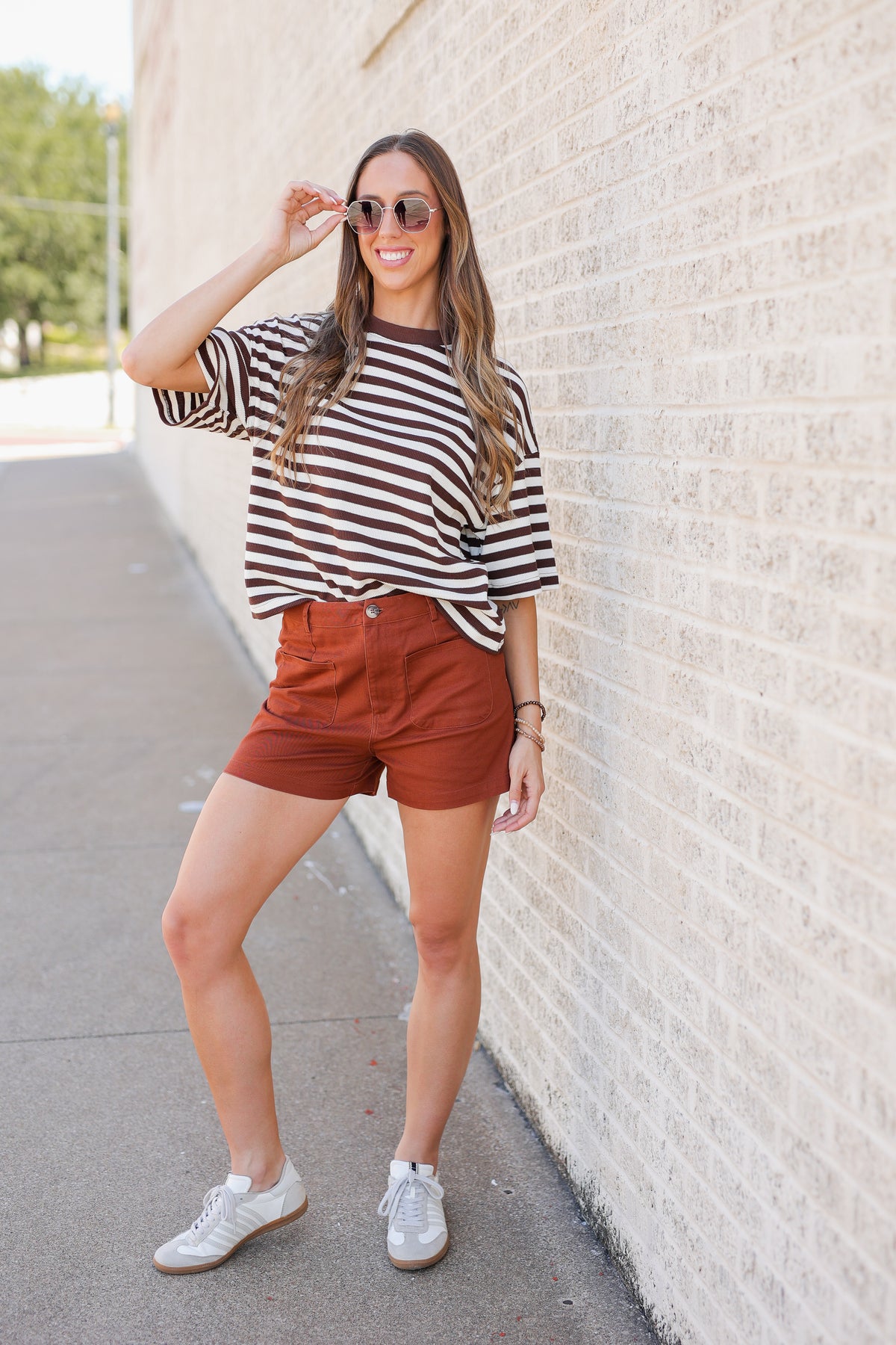A woman posing outdoors against a light brick wall, wearing a brown and white striped t-shirt, rust-colored shorts, and white sneakers.