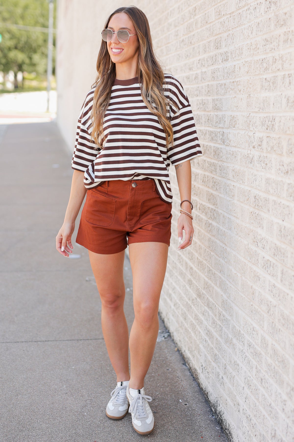 A woman stands outdoors against a light brick wall wearing a brown and white horizontally striped short-sleeve t-shirt, rust-brown shorts, silver sneakers, and round sunglasses.