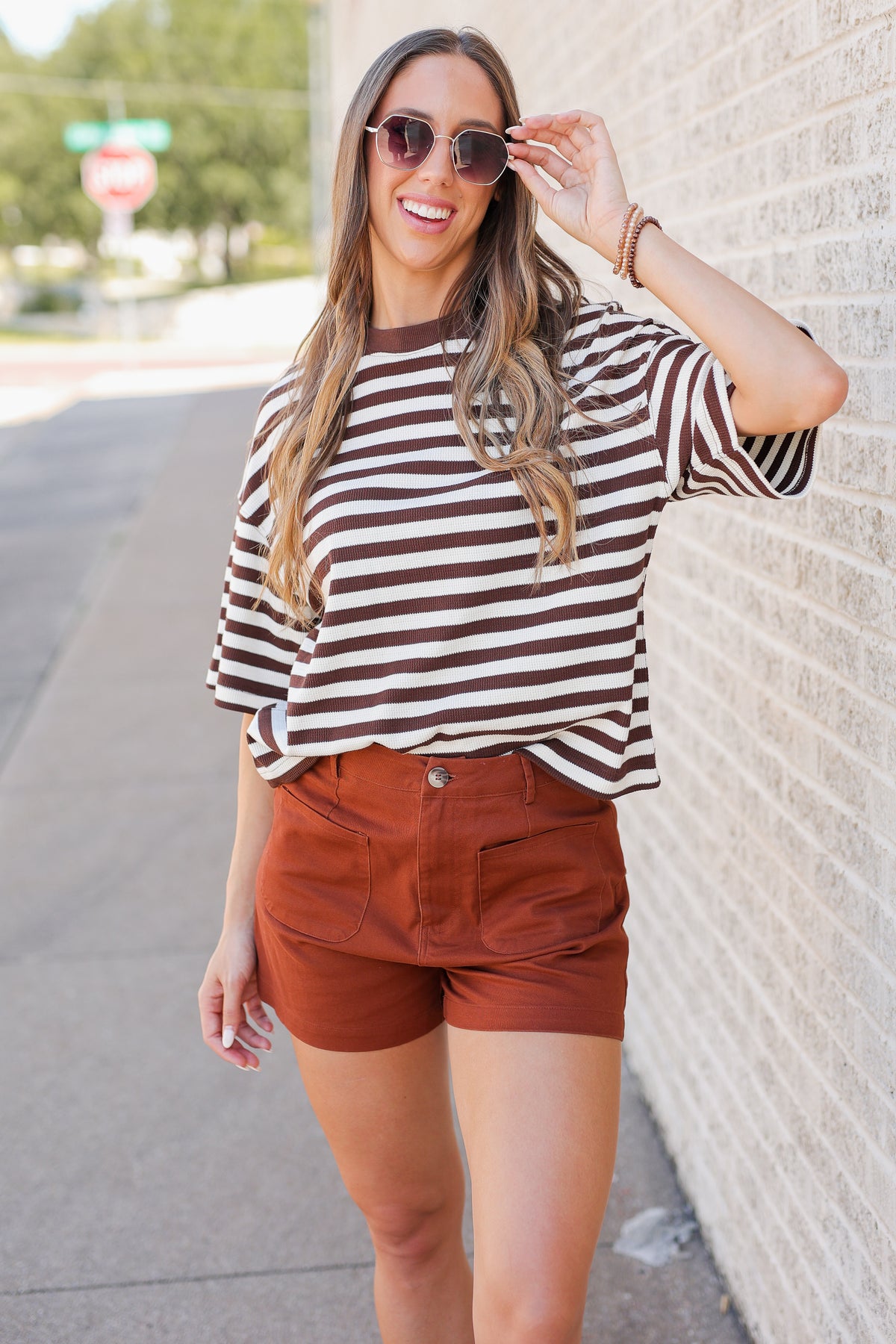 A woman wearing a brown and cream striped short-sleeve t-shirt and rust-colored shorts, posing outdoors against a light brick wall.