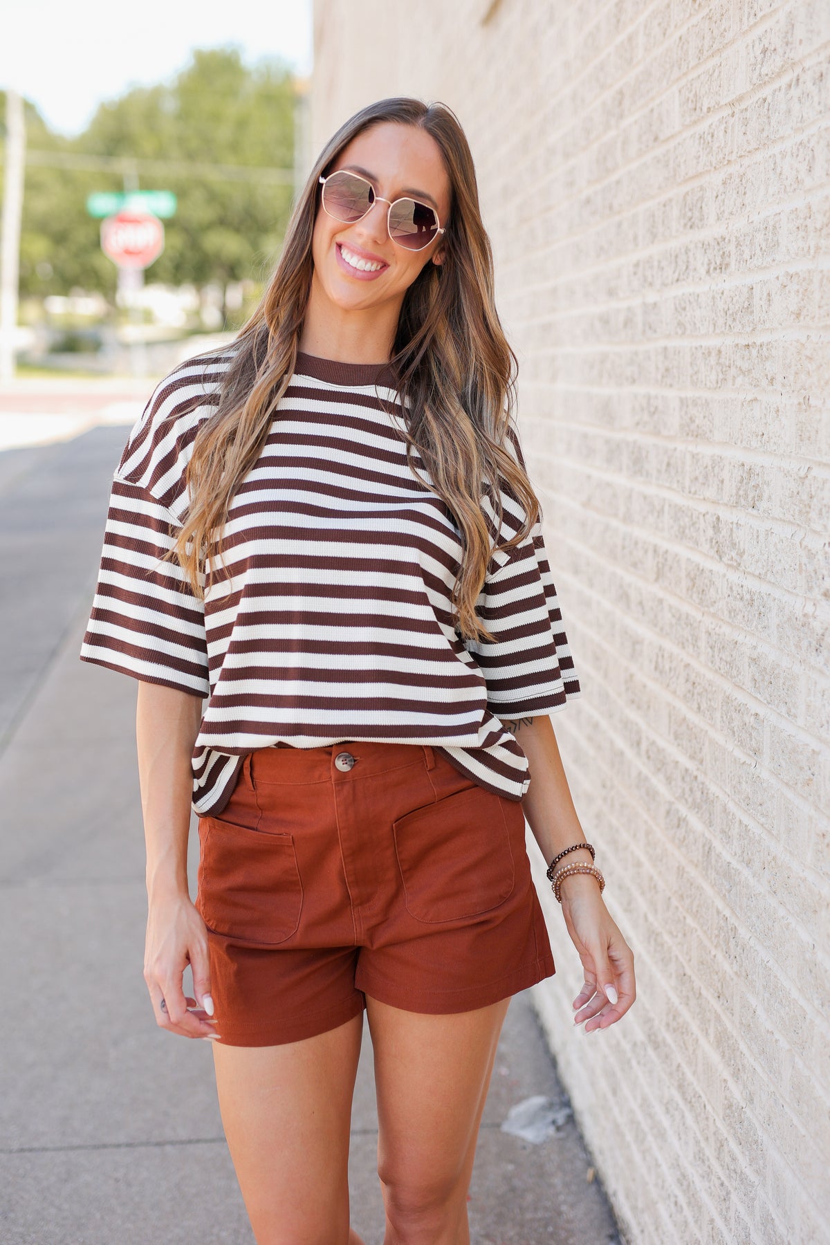 A woman wearing a brown and white striped short-sleeve t-shirt and rust-brown shorts, standing outdoors against a light brick wall.