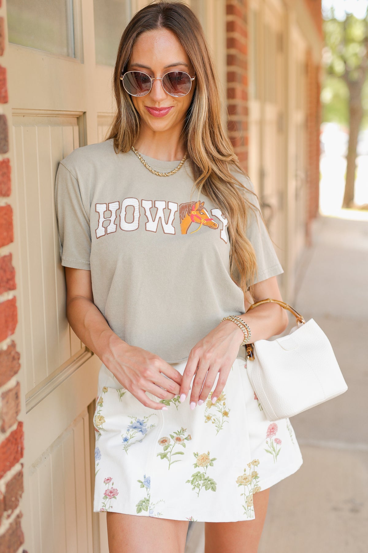 A woman wearing a light tan graphic t-shirt with 'HOWDY' and a peach slice graphic, paired with a white floral mini skirt and holding a white handbag.