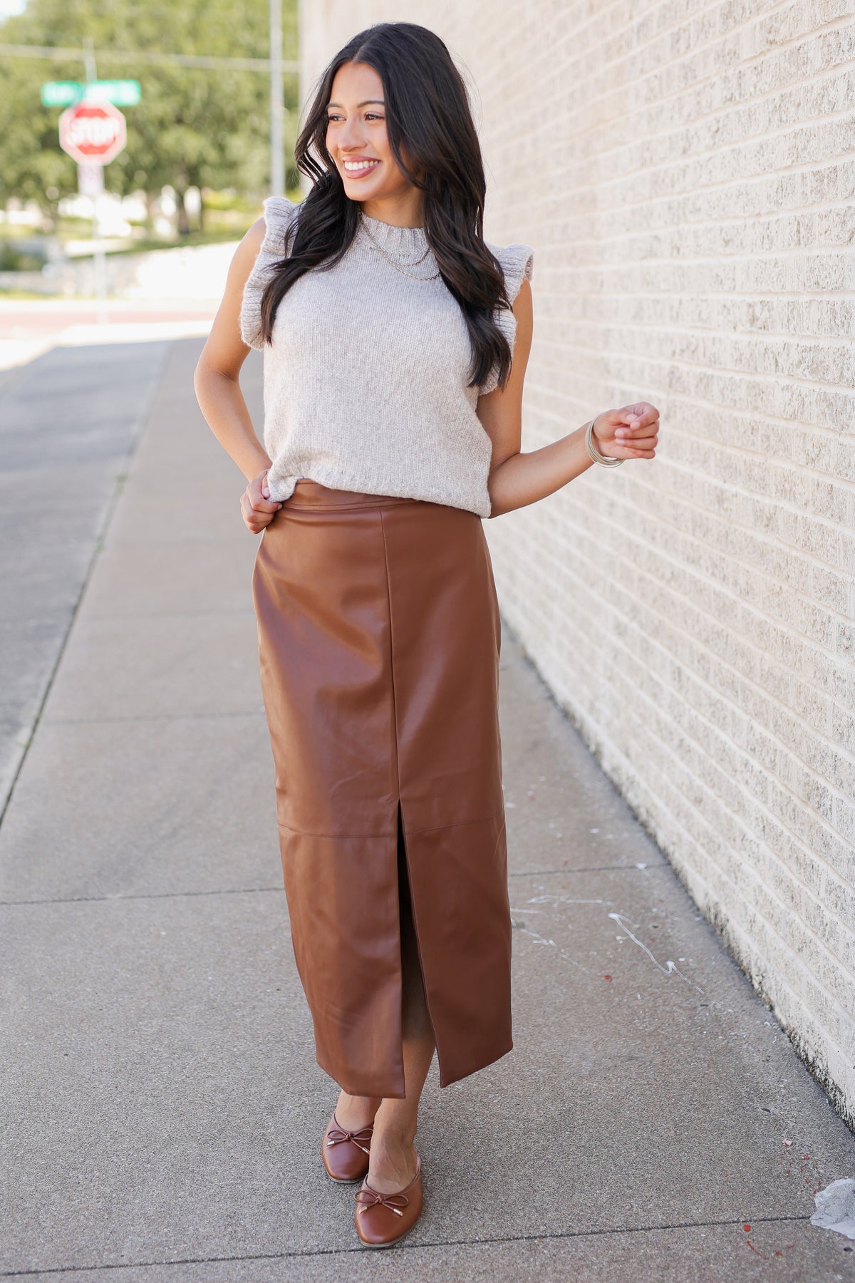 Model wearing a oatmeal colored sweater top with a cognac colored faux leather skirt standing outside.