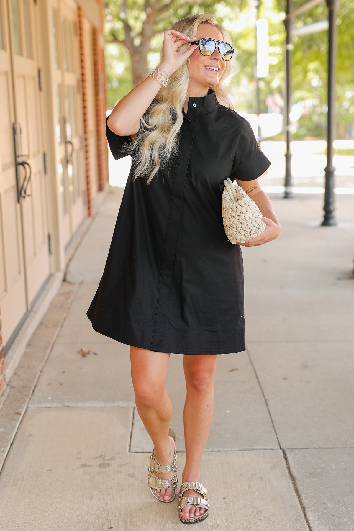 Woman in a black dress holding sunglasses and a clutch on a sidewalk.