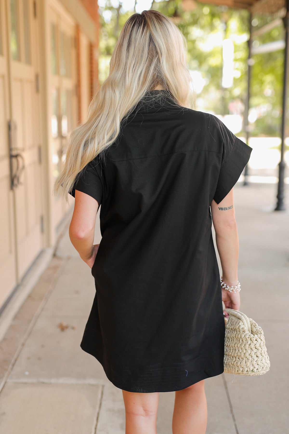 Person wearing a black dress holding a small woven bag on a sidewalk.