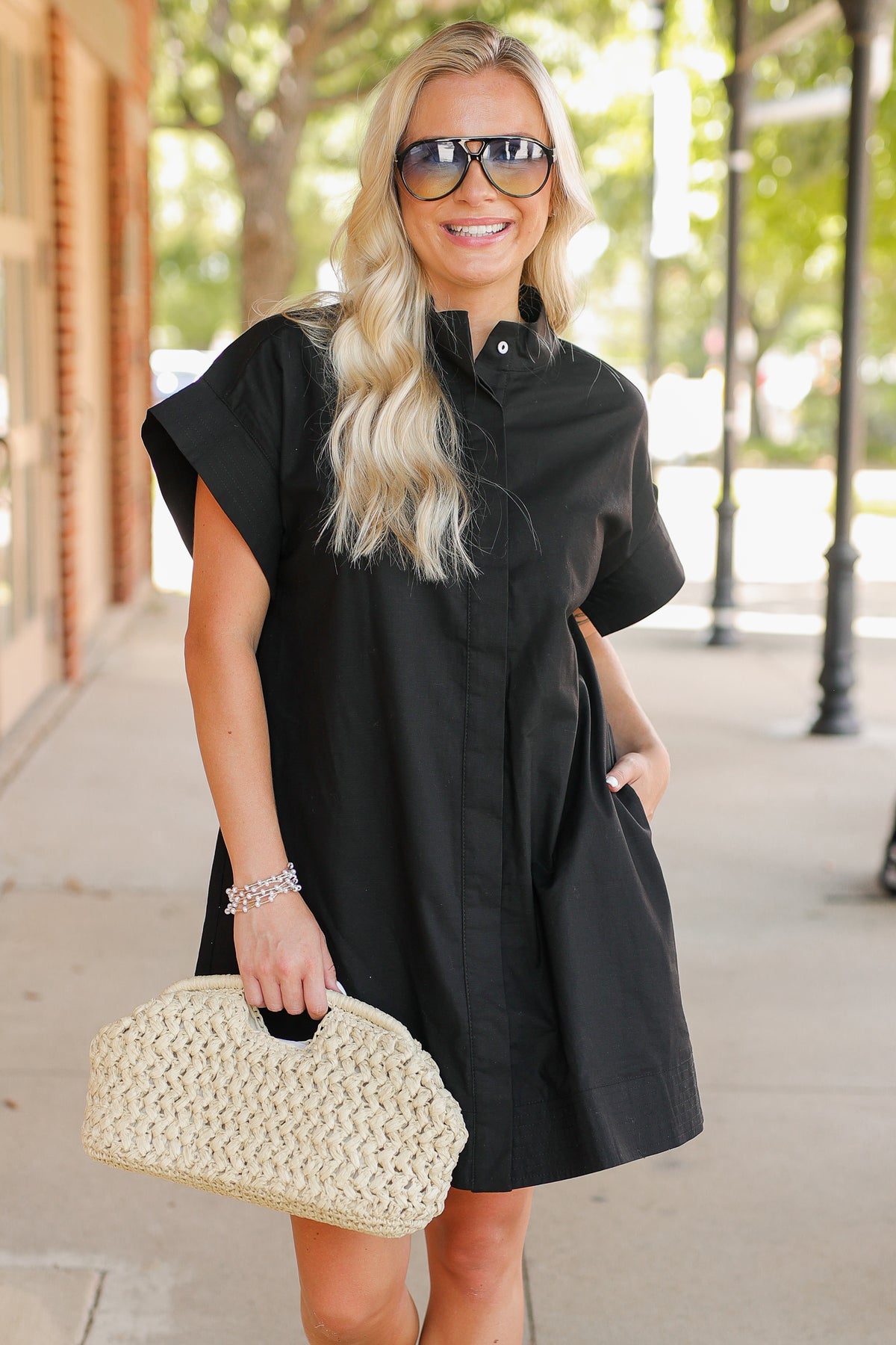 Woman wearing a black dress and sunglasses, holding a beige handbag on a sidewalk.