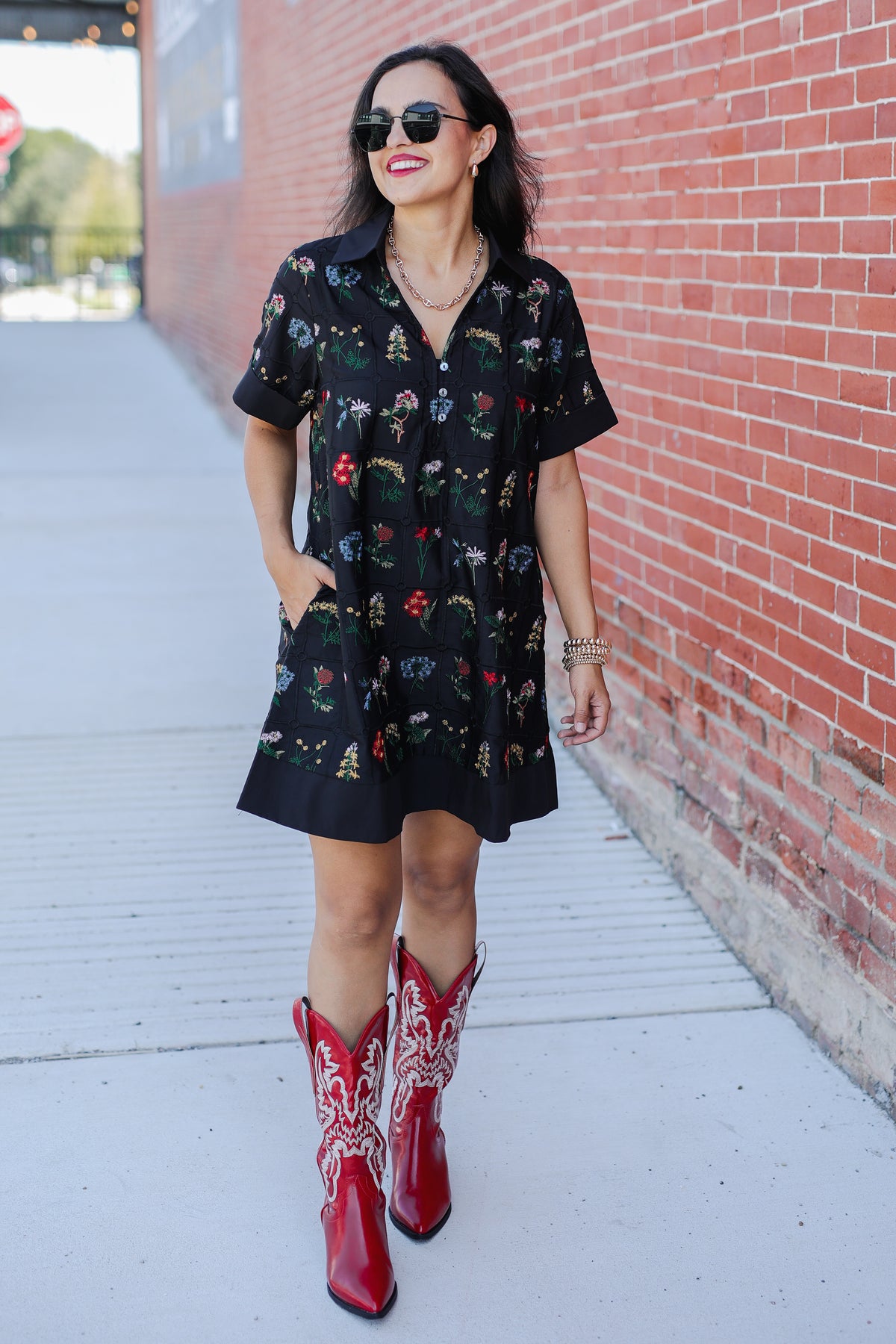 Woman wearing a black dress with colorful patterns and red cowboy boots against a brick wall.