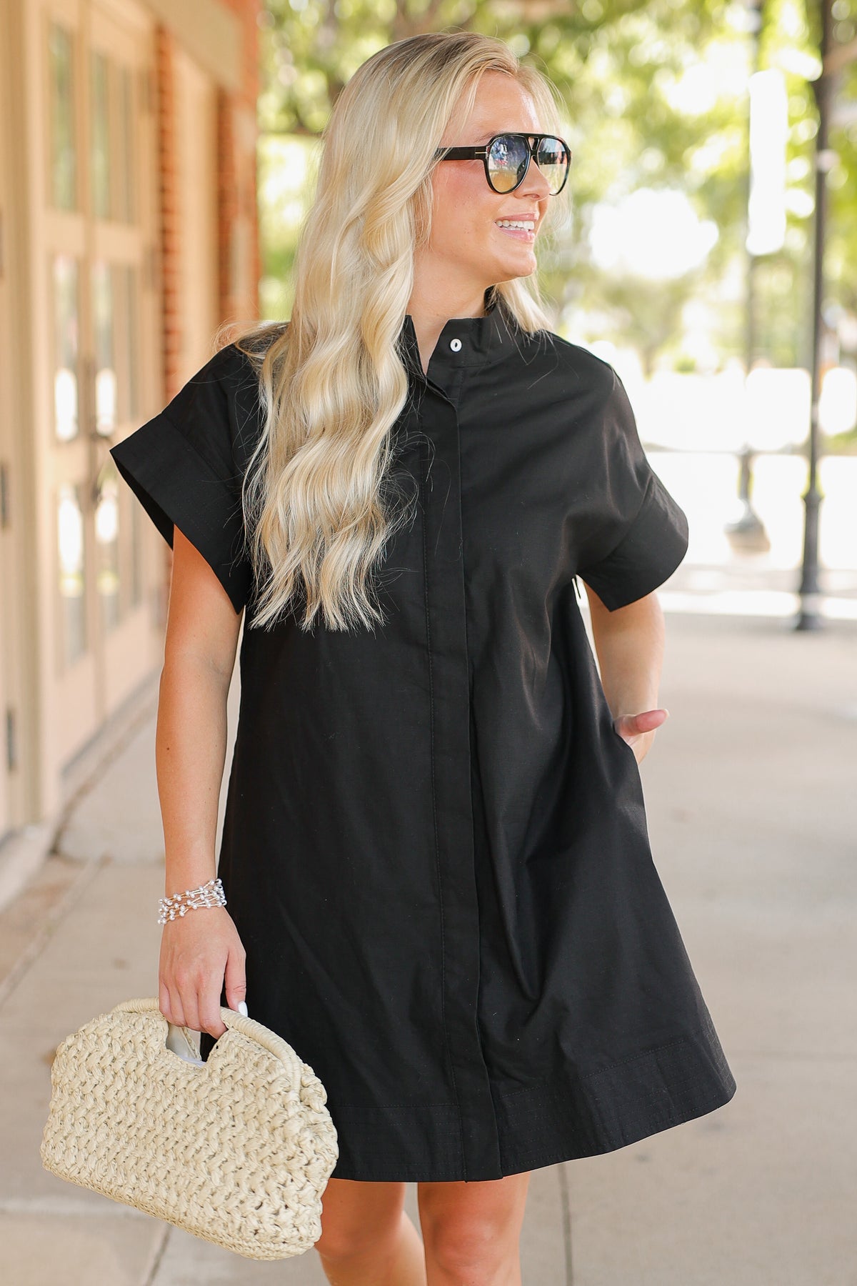Woman wearing a black dress with a straw handbag on a sidewalk.