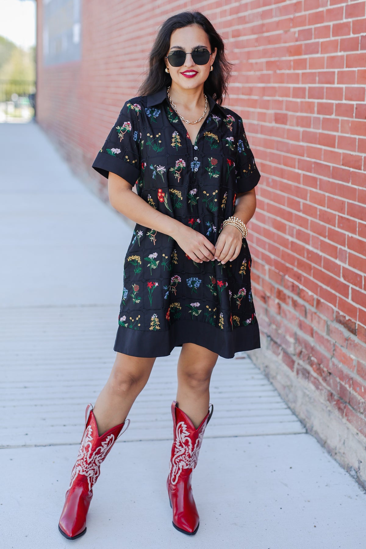 Woman wearing a black floral dress and red cowboy boots against a brick wall.