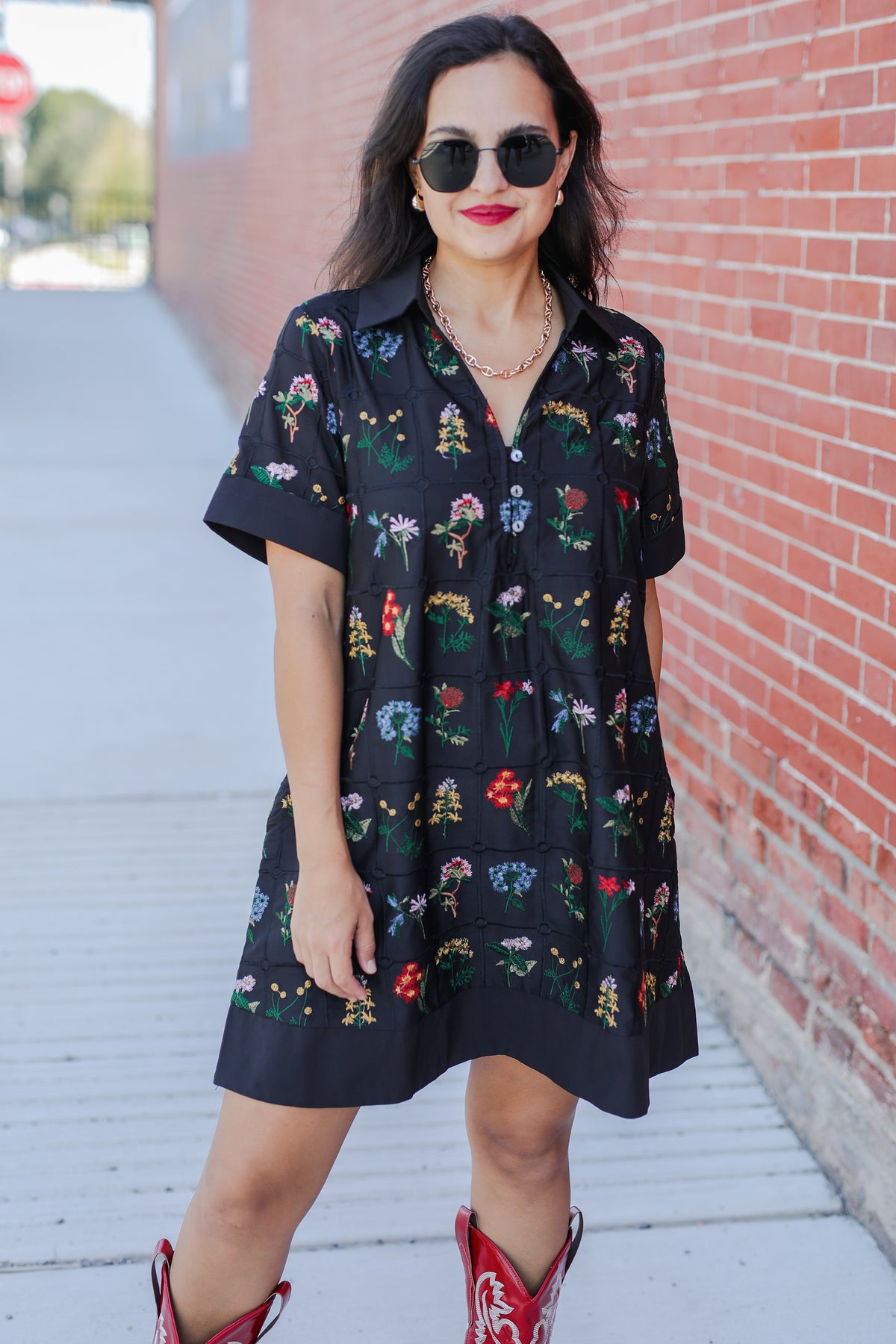 Woman wearing a black floral dress with red cowboy boots against a brick wall.