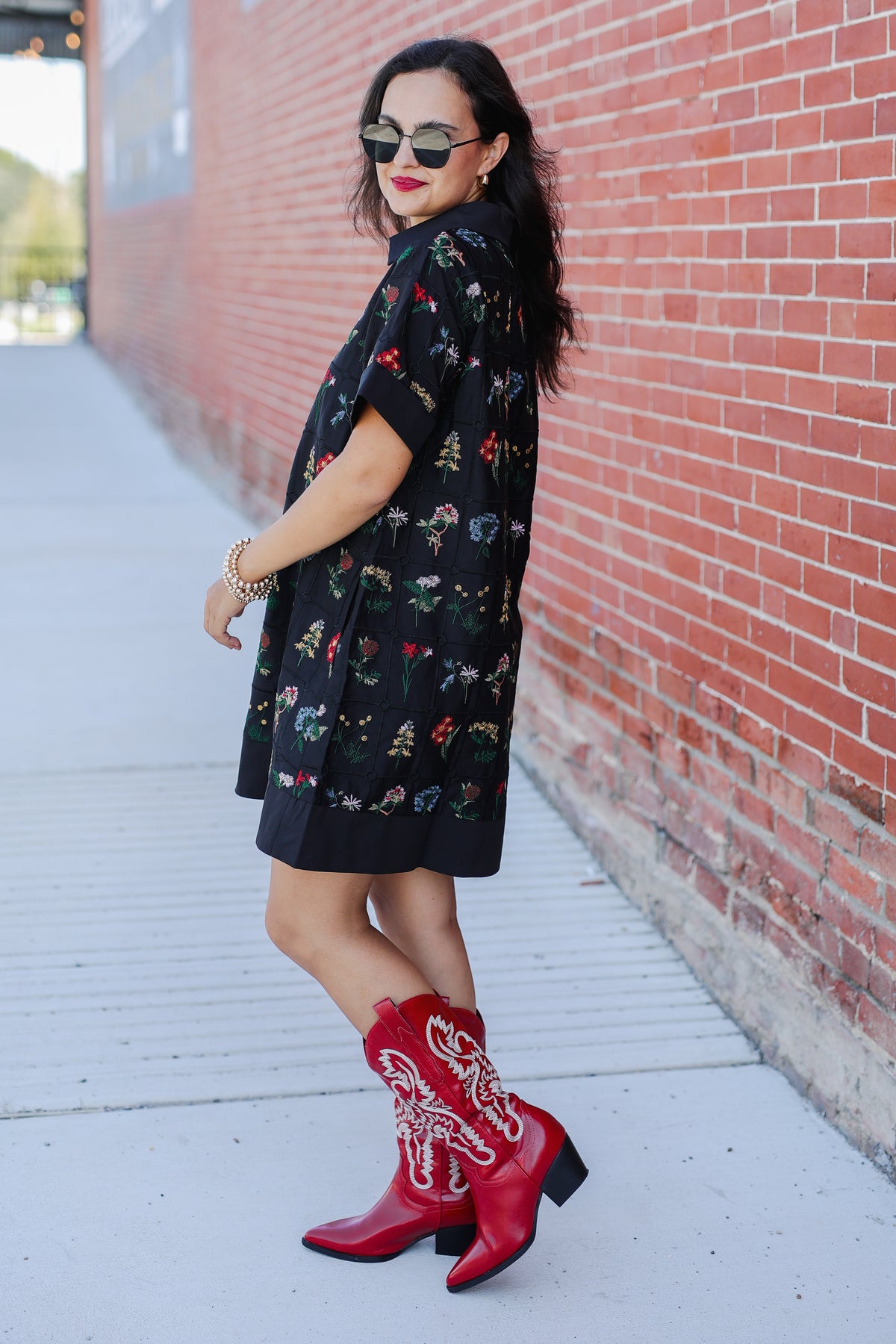 Woman wearing a black floral dress and red cowboy boots against a brick wall.