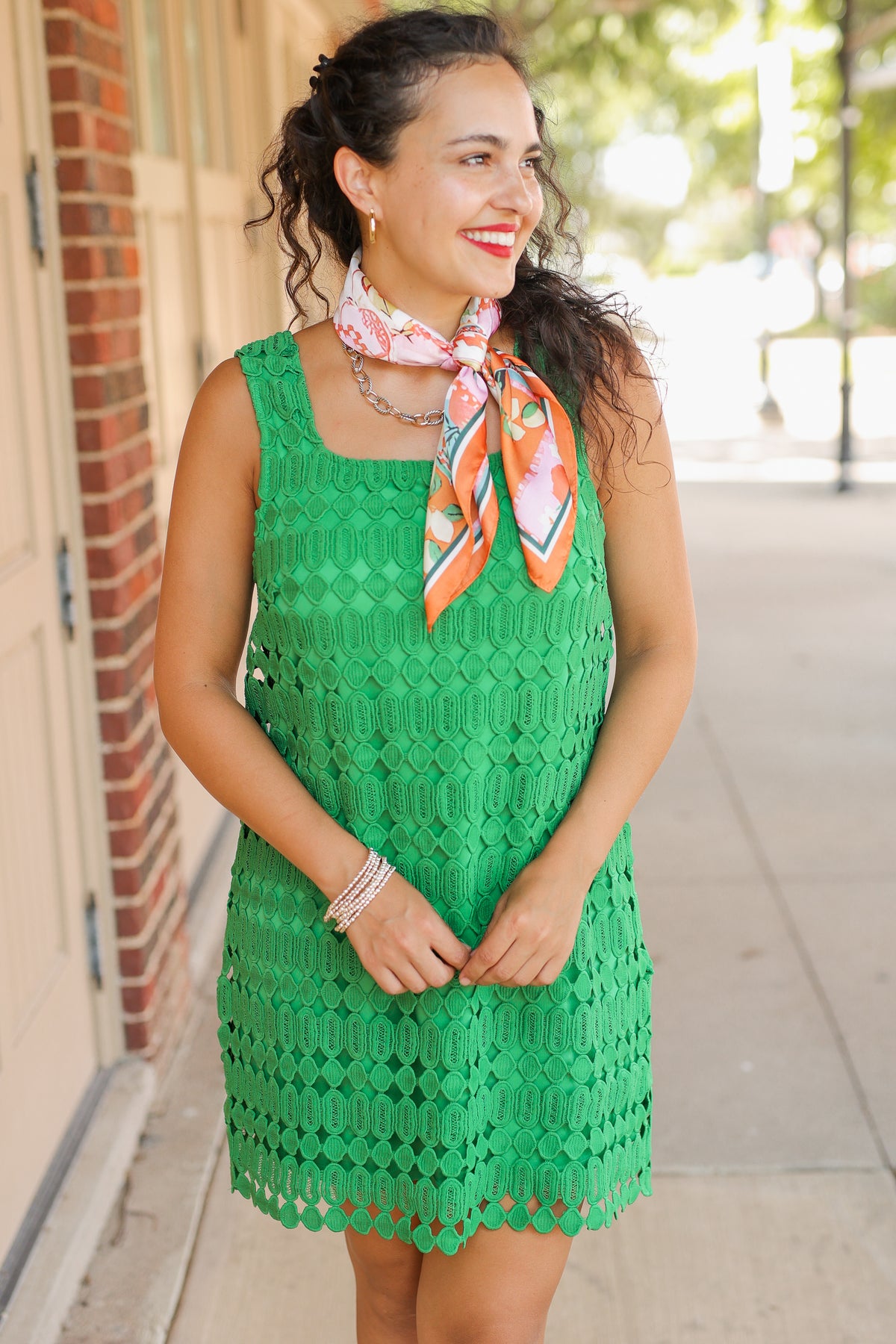 Woman wearing a green lace dress with an orange scarf, standing on a sidewalk.