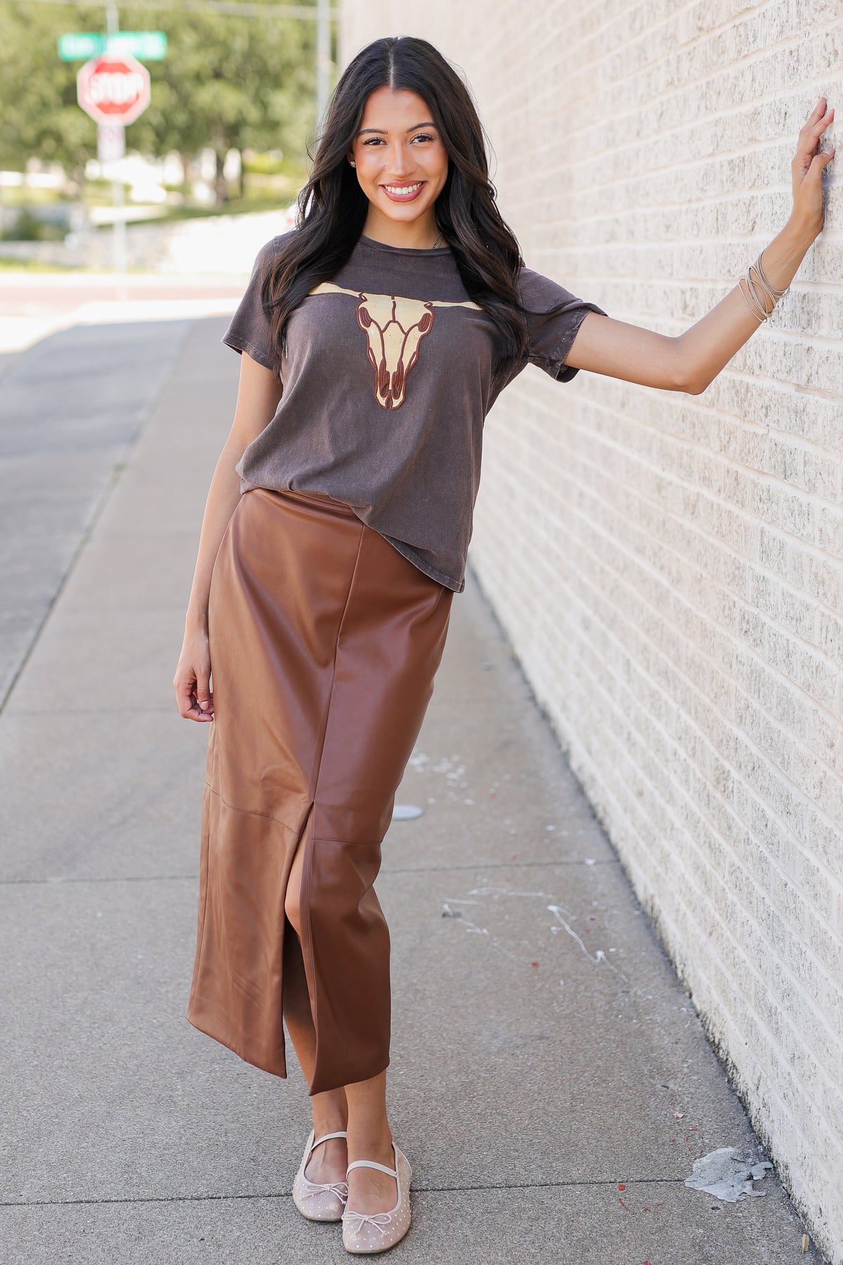 A woman stands on a sidewalk next to a white brick wall, wearing a dark brown graphic t-shirt with a cow print and a brown faux-leather midi skirt.