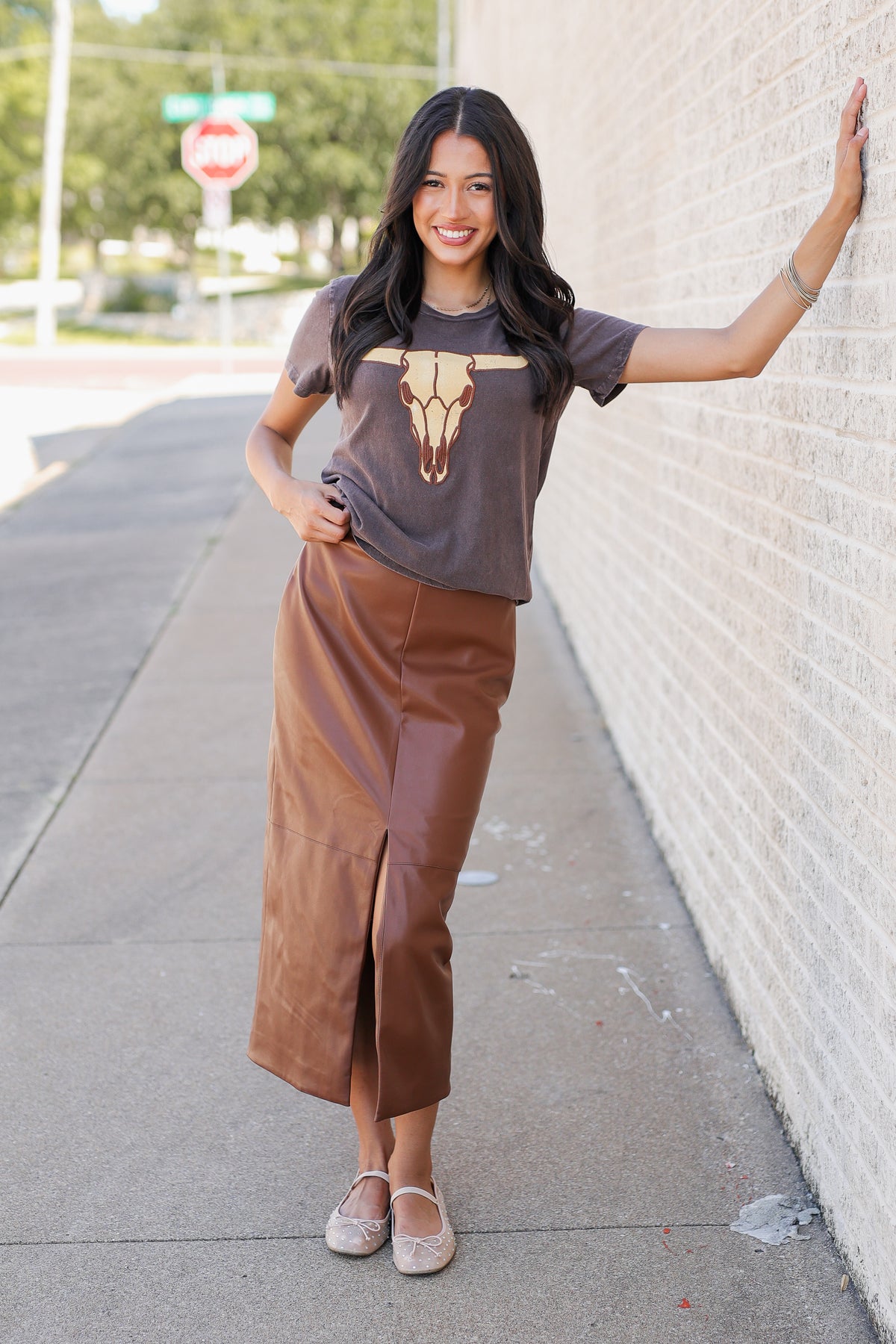 A woman stands on a sidewalk against a white brick wall, wearing a brown graphic t-shirt with a bull skull design and a matching brown faux-leather midi skirt.