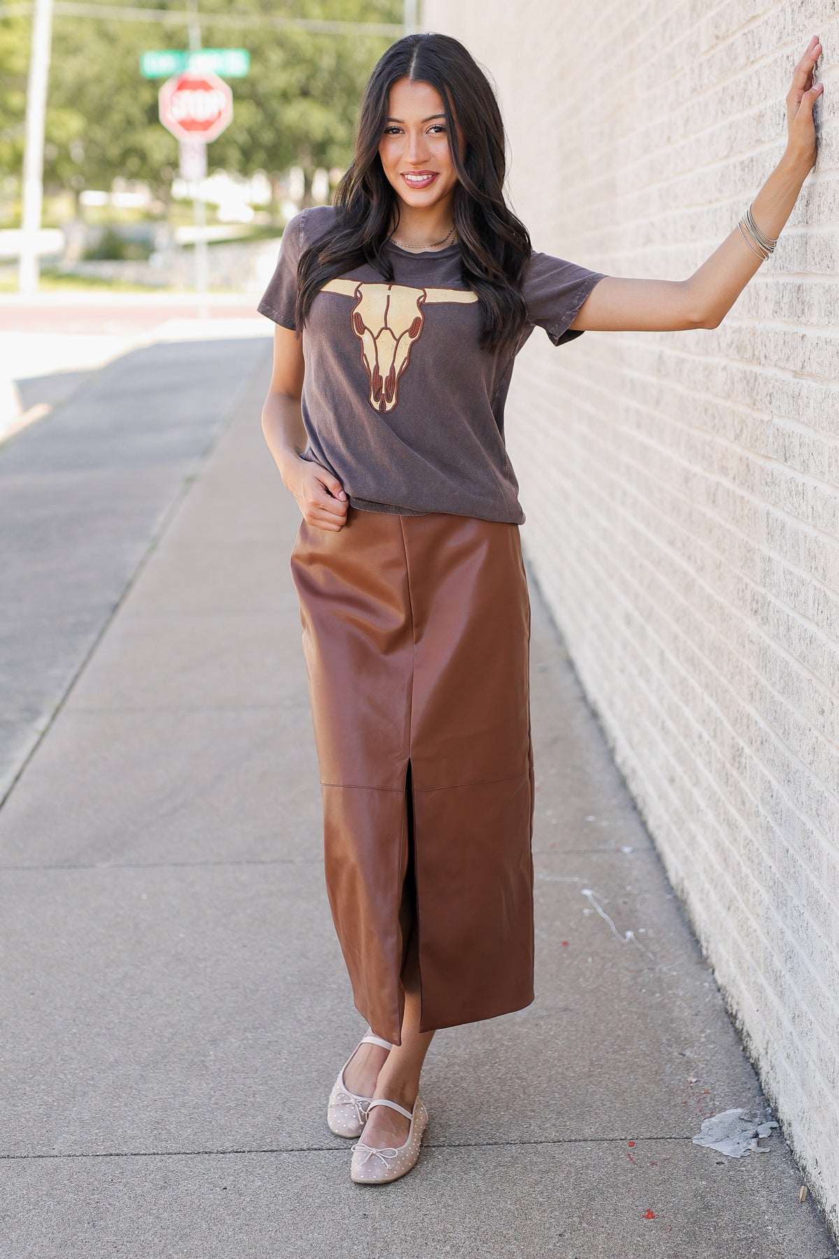 A woman stands on a sidewalk against a light brick wall, wearing a brown graphic t-shirt with a steer skull print and a brown faux-leather midi skirt with a front slit.