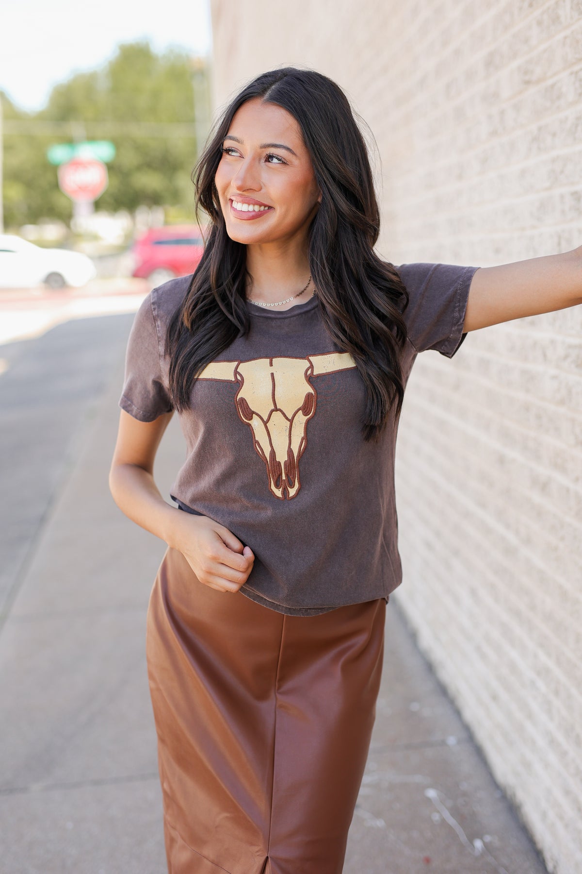 A woman stands on a sidewalk next to a light brick wall, wearing a dark brown graphic t-shirt with a longhorn skull design and a brown faux-leather skirt.