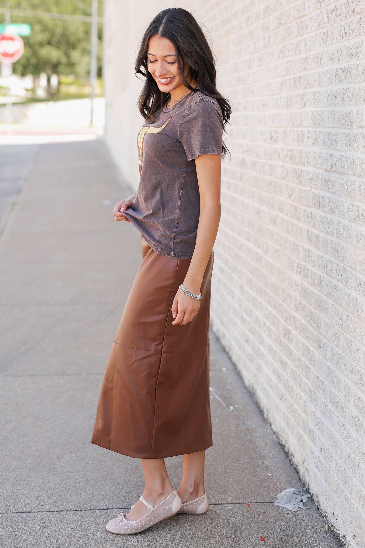 A woman stands on a sidewalk beside a white brick wall, wearing a short-sleeve taupe-brown top and a matching brown midi skirt with light pink ballet flats.