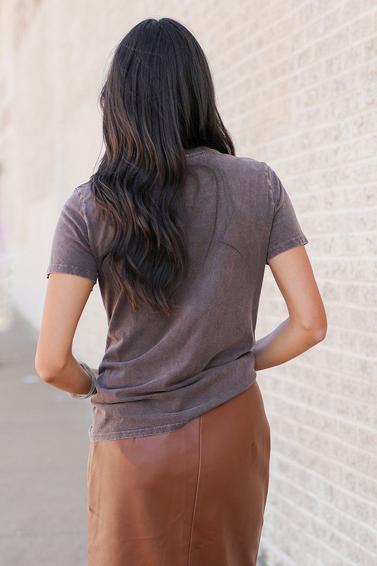 A woman viewed from behind wearing a short-sleeve, crew-neck t-shirt in a washed, heathered taupe-brown color, paired with a brown skirt, standing against a light-colored brick wall.