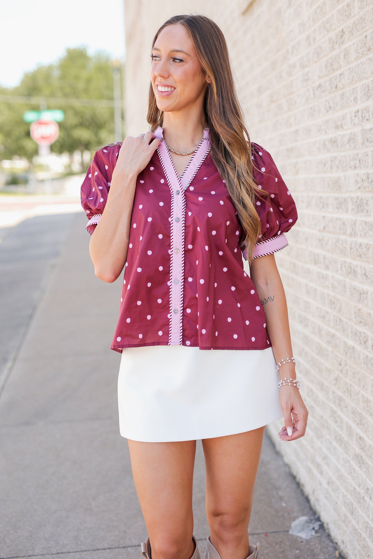 A woman wearing a burgundy short-sleeve button-up blouse with white polka dots and pink trim, paired with a white mini skirt, standing outdoors next to a light-colored brick wall.
