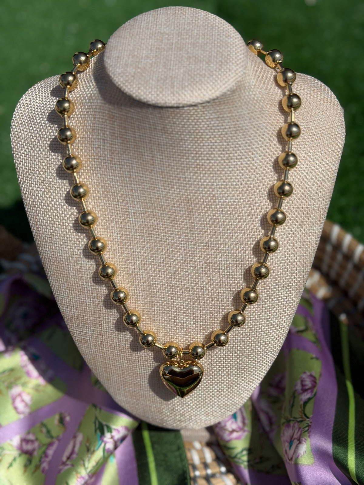 Gold beaded necklace with a heart pendant on a beige mannequin bust against a green and purple floral background.