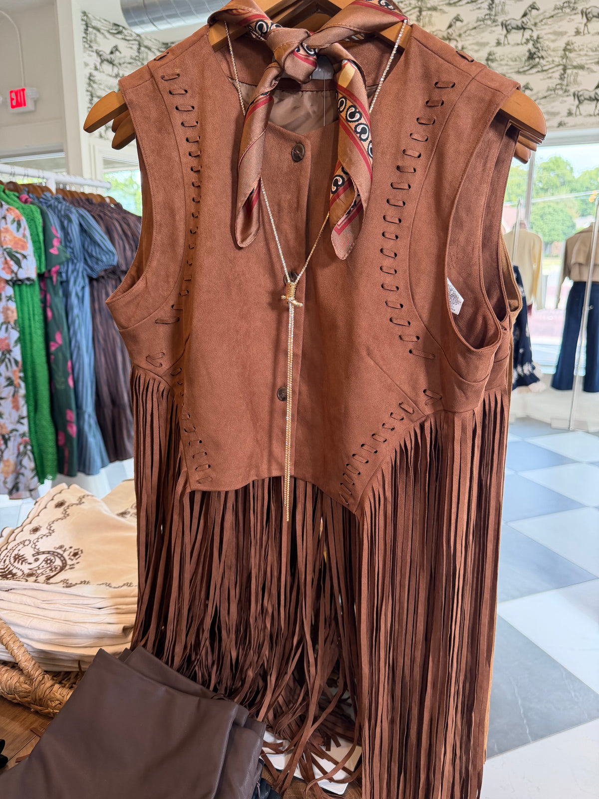 A brown suede fringe vest with decorative cutouts and a matching neck scarf, displayed on a clothing rack inside a retail store.