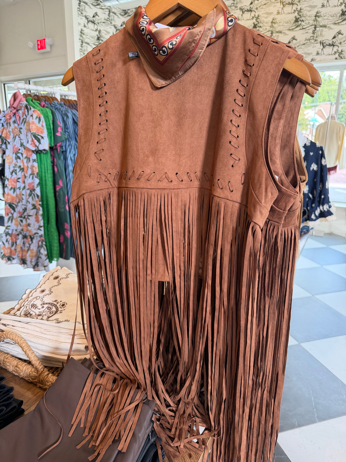 A brown suede fringe vest with decorative cutouts and a matching patterned bandana collar, displayed on a wooden hanger inside a clothing store.